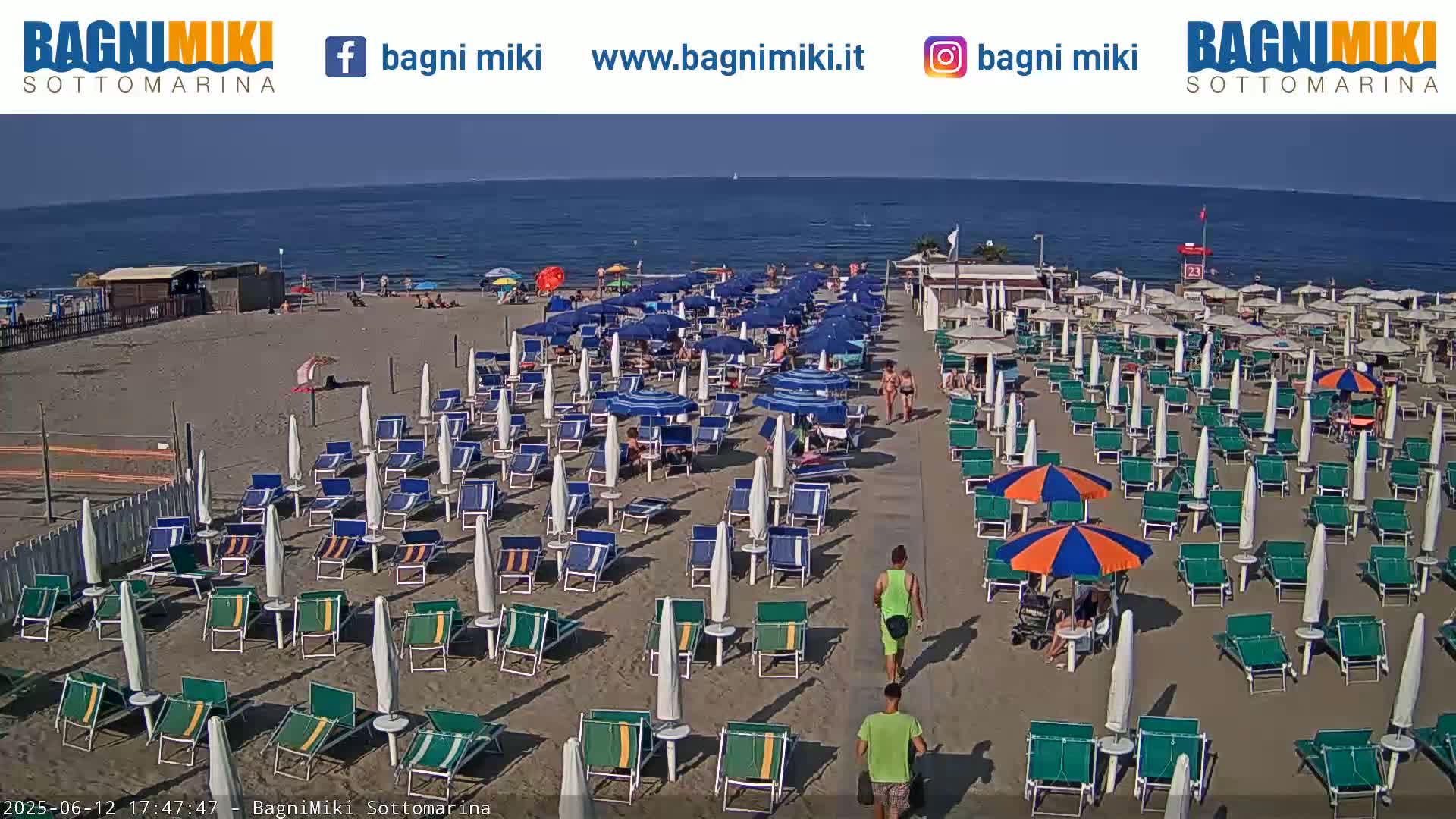 A sandy beach with numerous beach umbrellas and lounge chairs is shown, next to a calm sea on a sunny day.