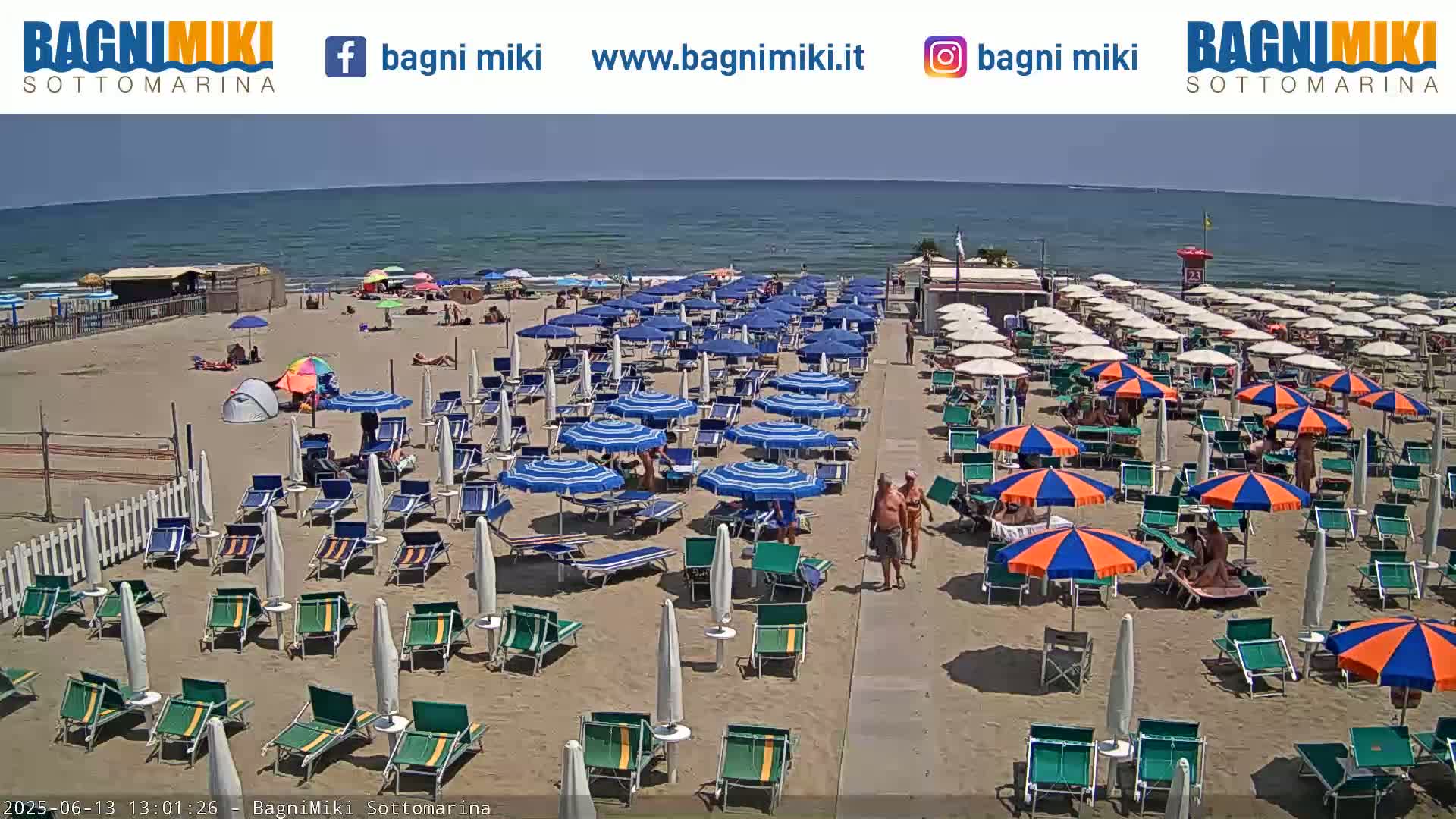 A sandy beach with many blue and orange umbrellas, beach chairs, and people enjoying a sunny day by a calm ocean.