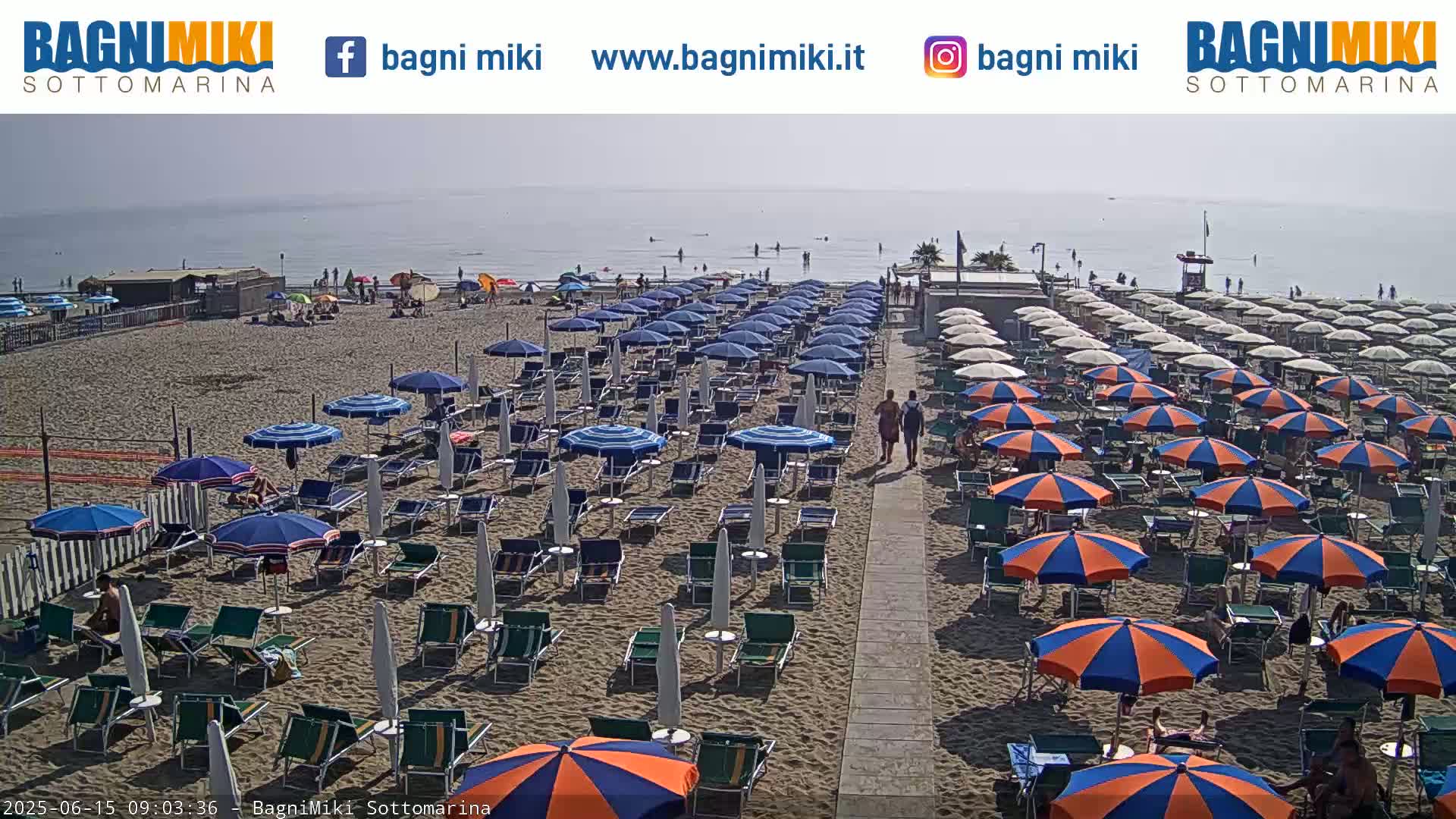 A sandy beach with numerous blue and orange umbrellas and lounge chairs is situated next to a calm, hazy ocean.