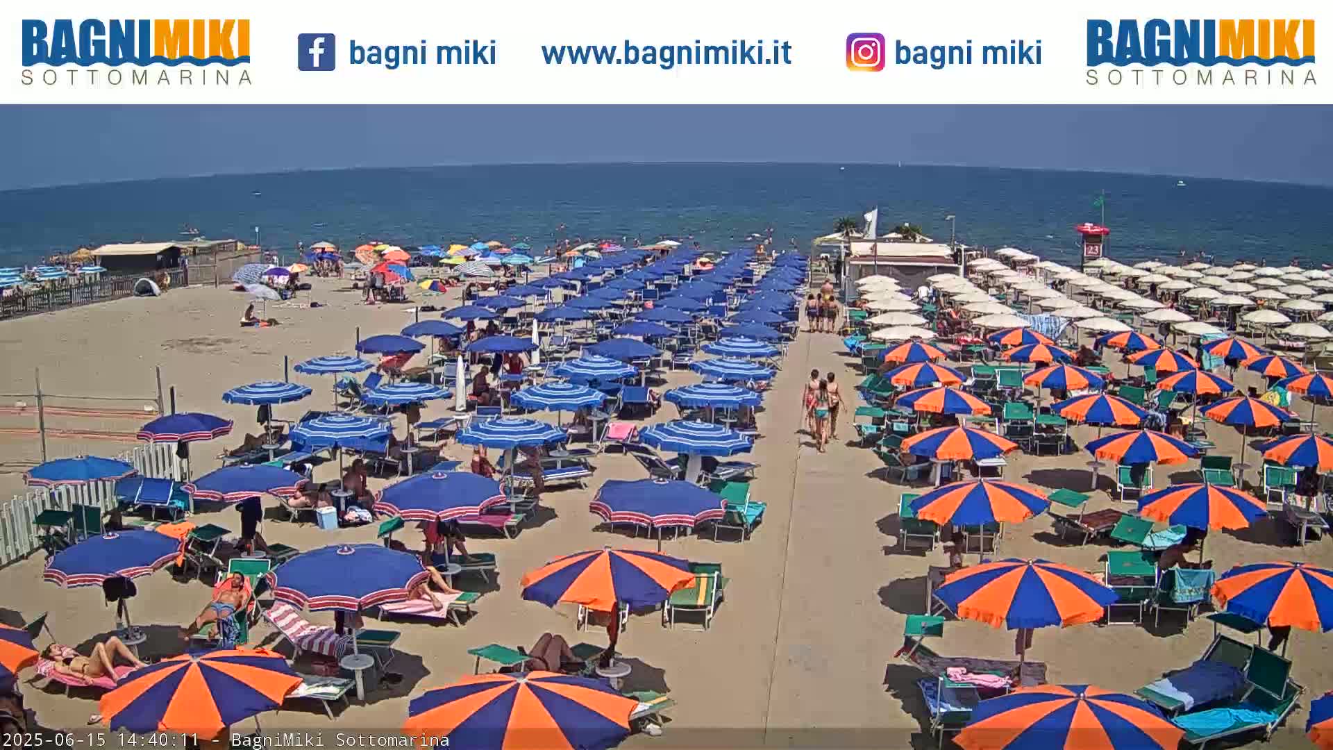 A sandy beach with many blue and orange umbrellas and beachgoers under a clear, sunny sky and calm ocean.