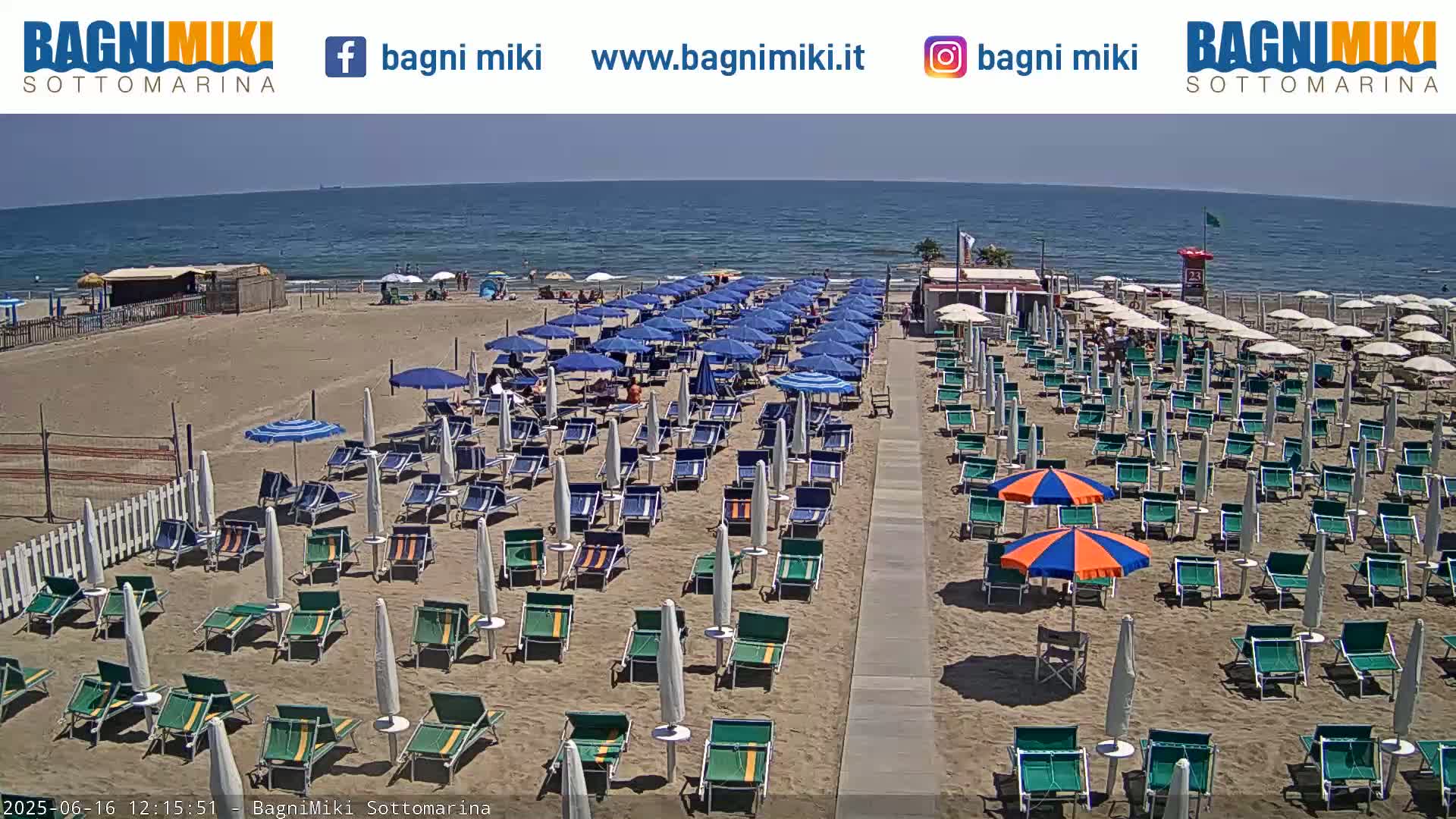 A sandy beach with numerous rows of blue and green beach chairs and umbrellas under a sunny sky next to a calm ocean.