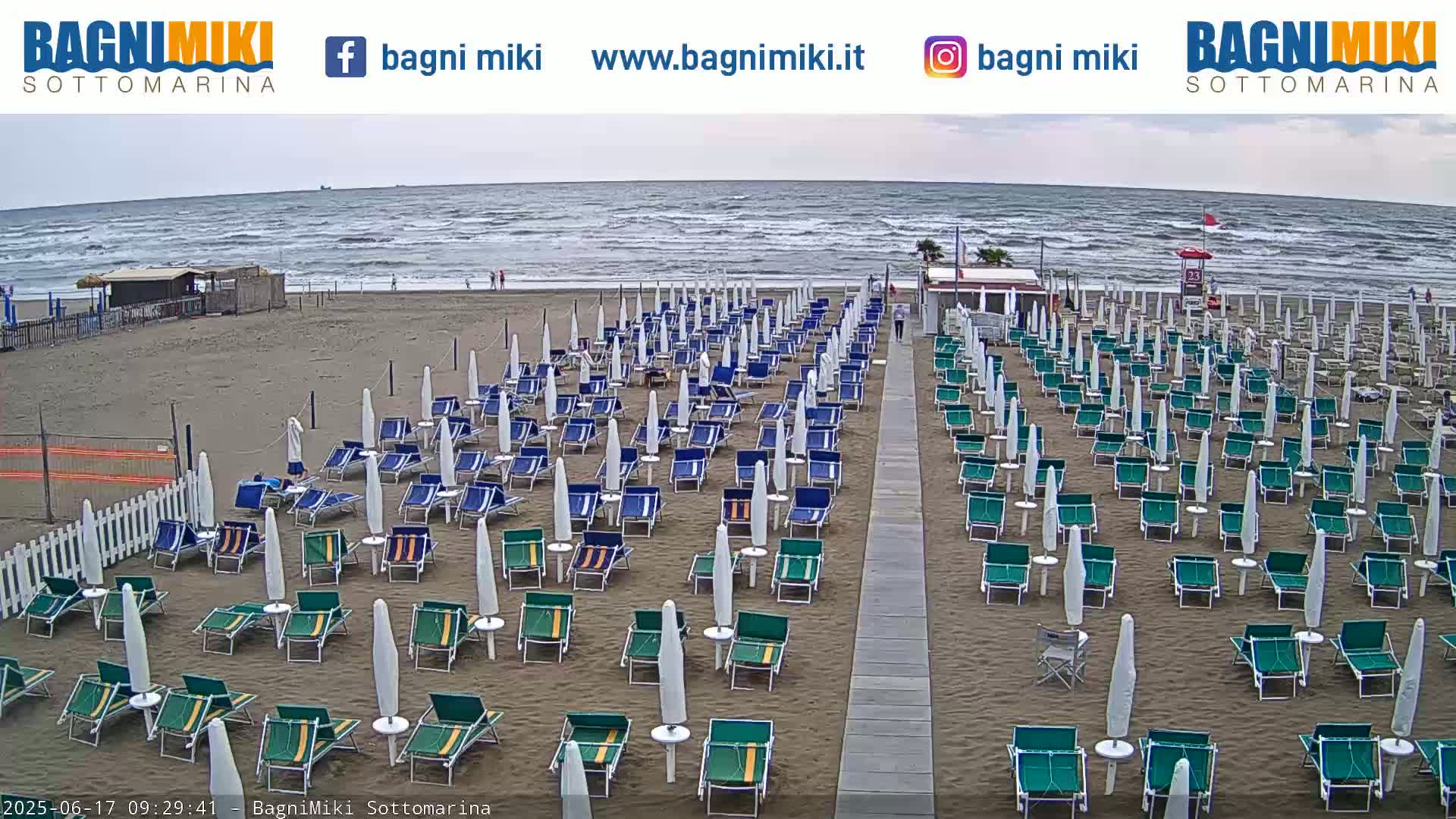 Rows of beach umbrellas and lounge chairs sit on a sandy beach overlooking a wavy, gray sea under an overcast sky.