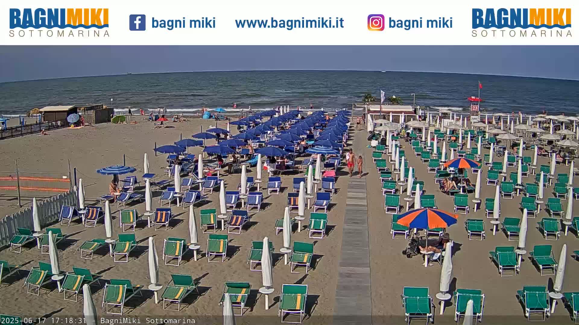 A sandy beach with numerous rows of beach umbrellas and lounge chairs is shown under a clear sky, next to a calm ocean.
