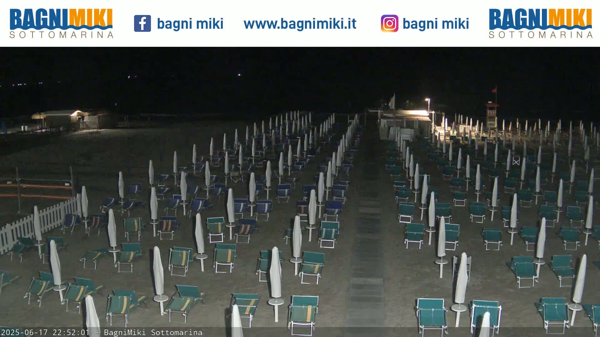 A nighttime beach scene showing numerous closed beach umbrellas and lounge chairs arranged in rows on a sandy beach.