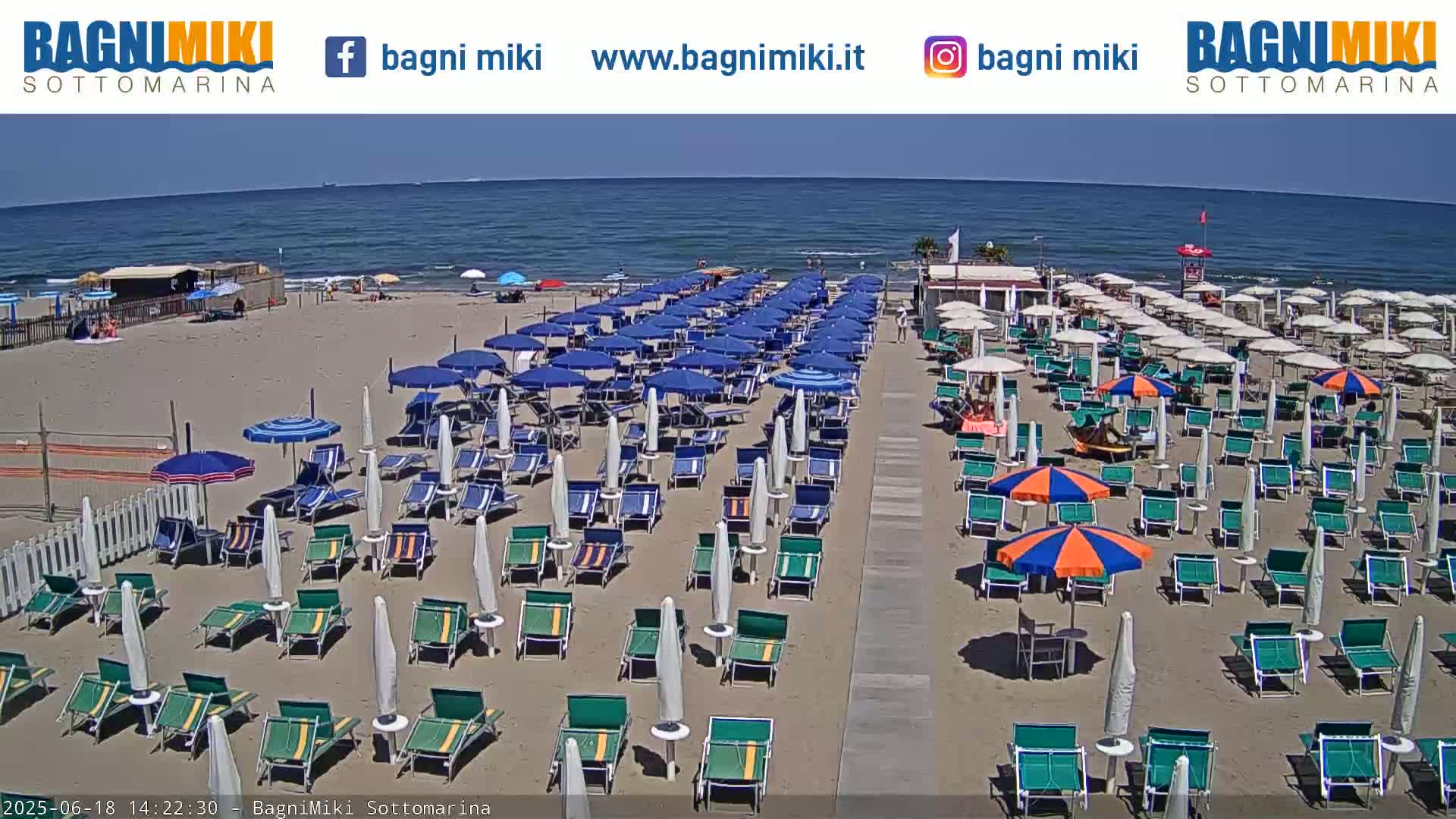 A sandy beach with numerous blue and orange beach umbrellas and lounge chairs is situated next to a calm ocean under a clear, sunny sky.