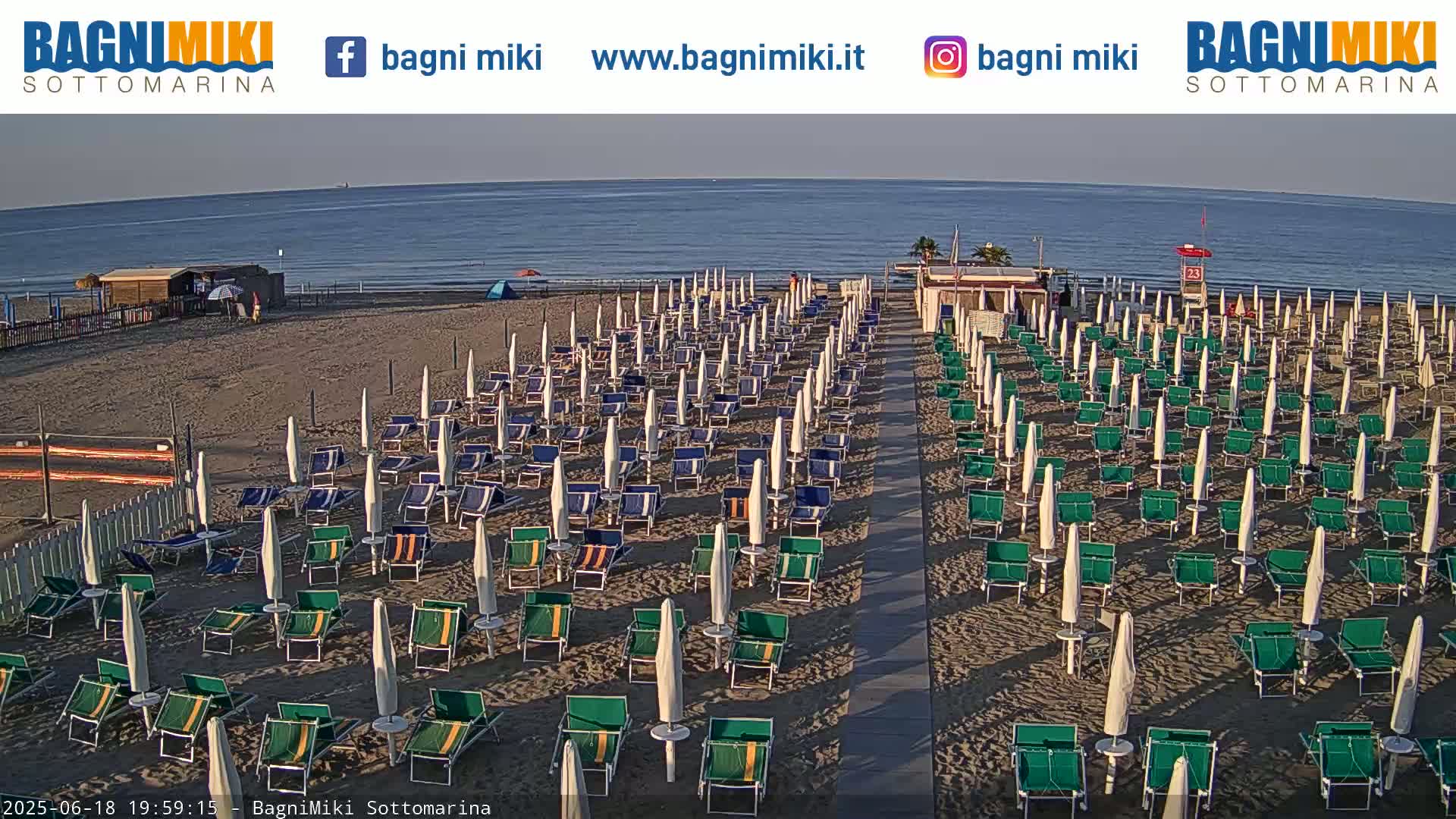 A sandy beach with numerous rows of closed beach umbrellas and lounge chairs, under a clear, sunny sky next to a calm ocean.