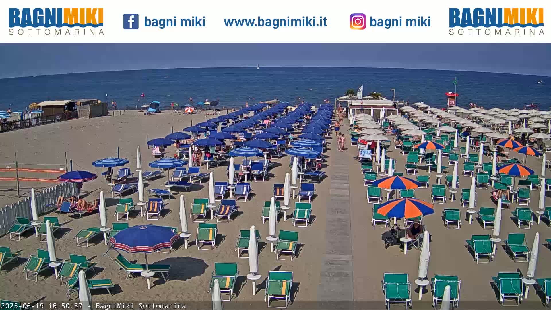 A sandy beach with numerous umbrellas and lounge chairs is crowded with people under a clear, sunny sky next to a calm blue ocean.