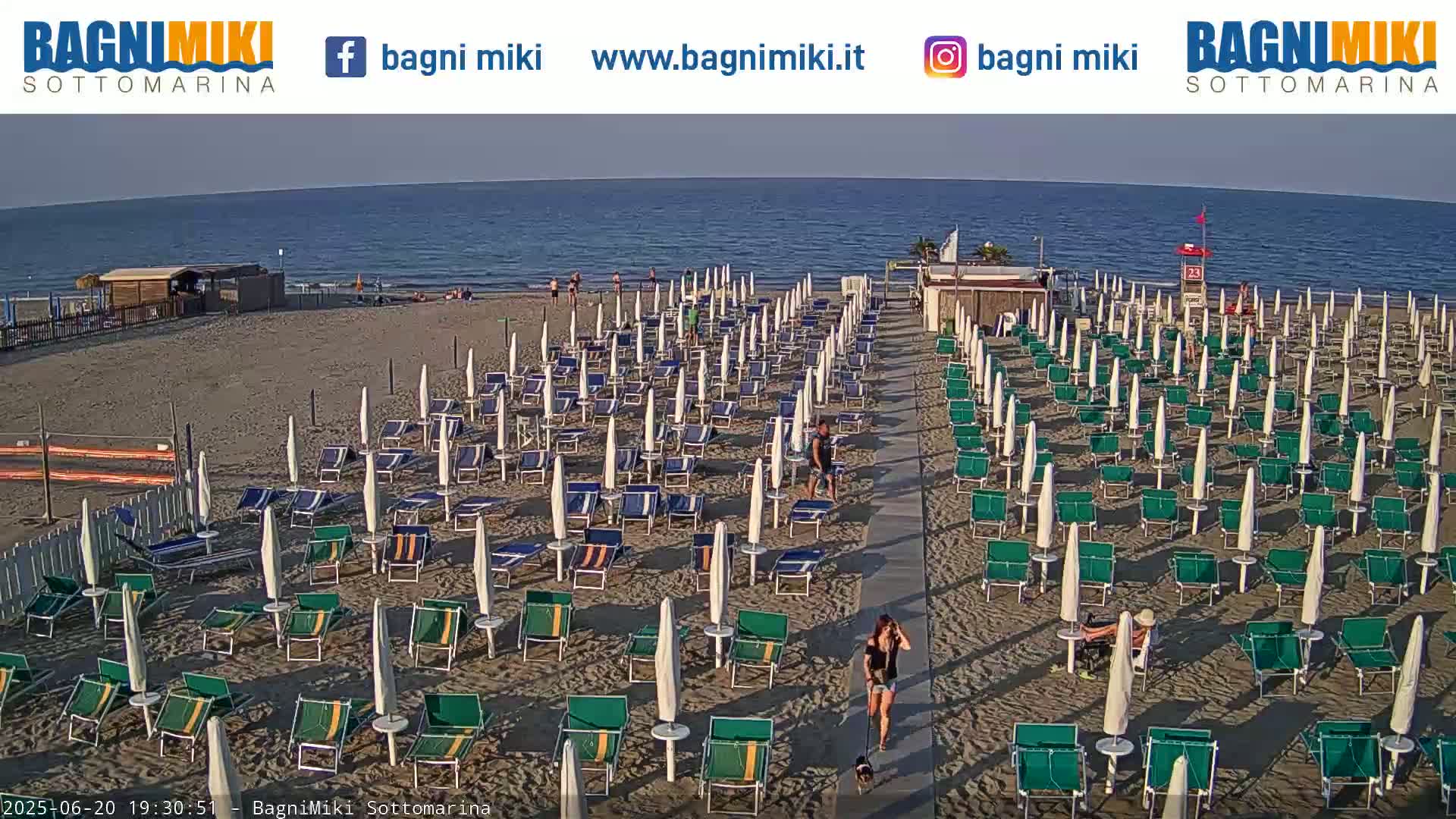 A sandy beach with numerous beach umbrellas and lounge chairs under a clear sky, next to a calm ocean.