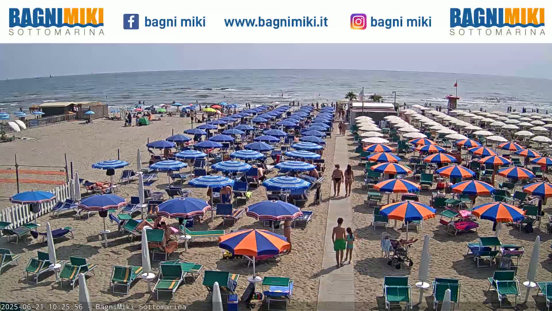 A sunny beach with numerous blue and orange umbrellas, beach chairs, and people enjoying a day at the seaside.
