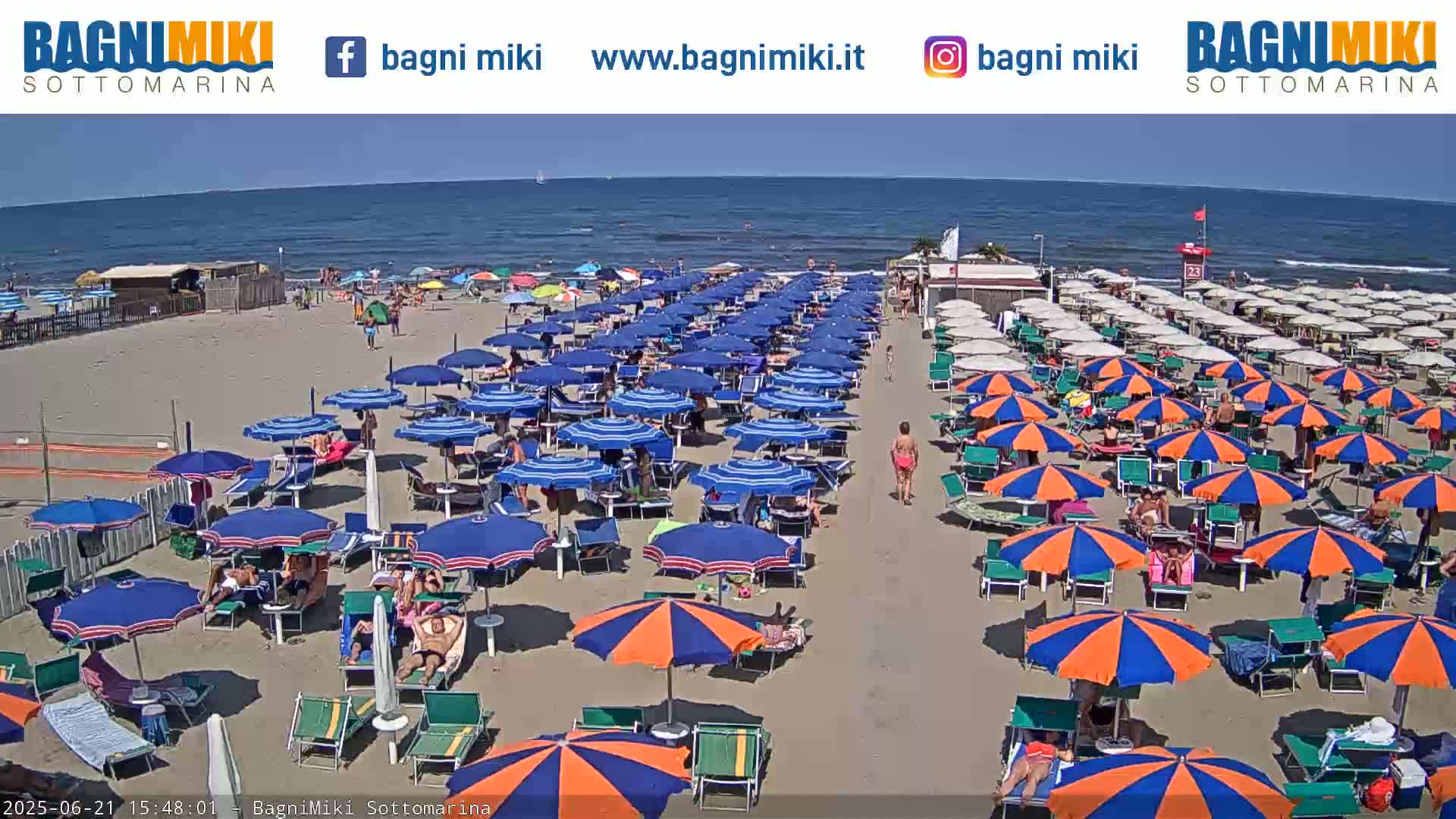 A sunny beach scene shows many blue and orange umbrellas and beach chairs lining the sandy shore, with a calm blue ocean in the background.