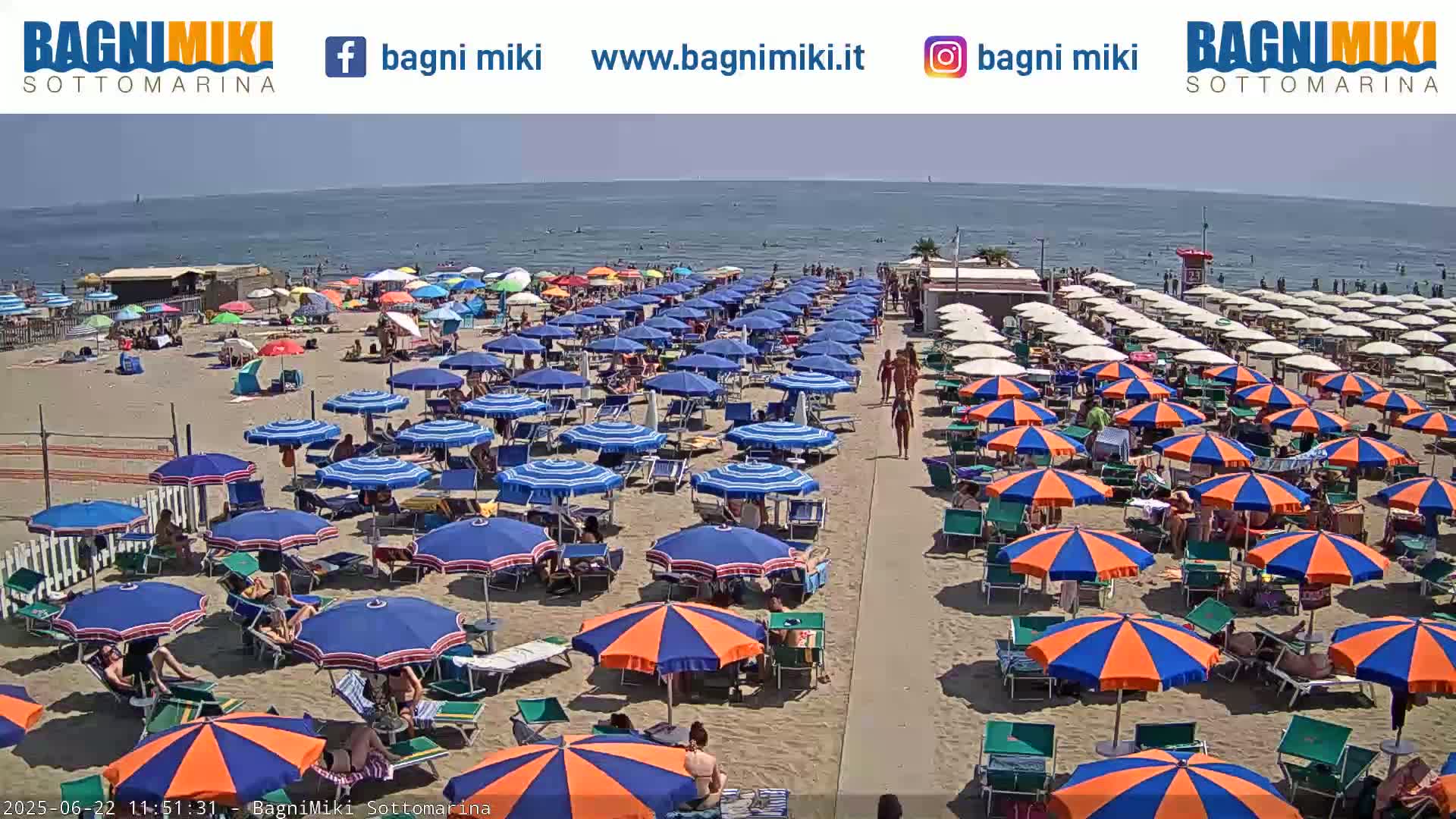 A sandy beach is crowded with numerous blue and orange umbrellas under a sunny sky, and the calm sea is visible in the background.