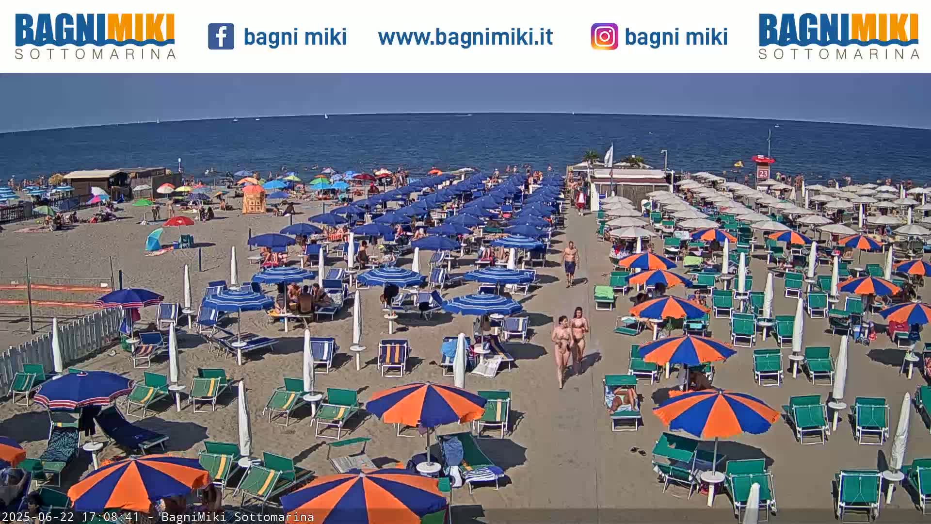 A sandy beach with numerous blue and orange umbrellas, beach chairs, and people, under a sunny sky, next to a calm sea.