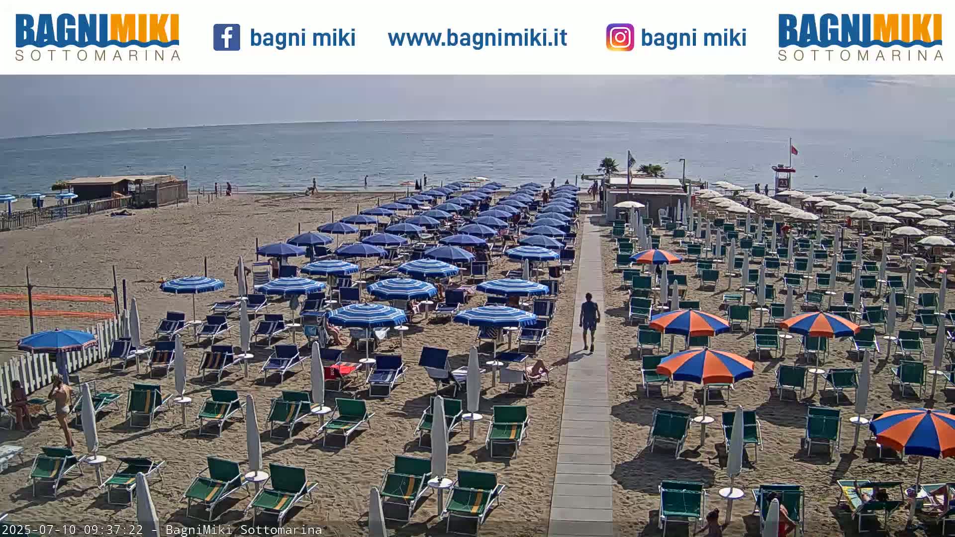 A sandy beach with numerous blue and orange umbrellas and lounge chairs is shown under a sunny sky, adjacent to a calm ocean.