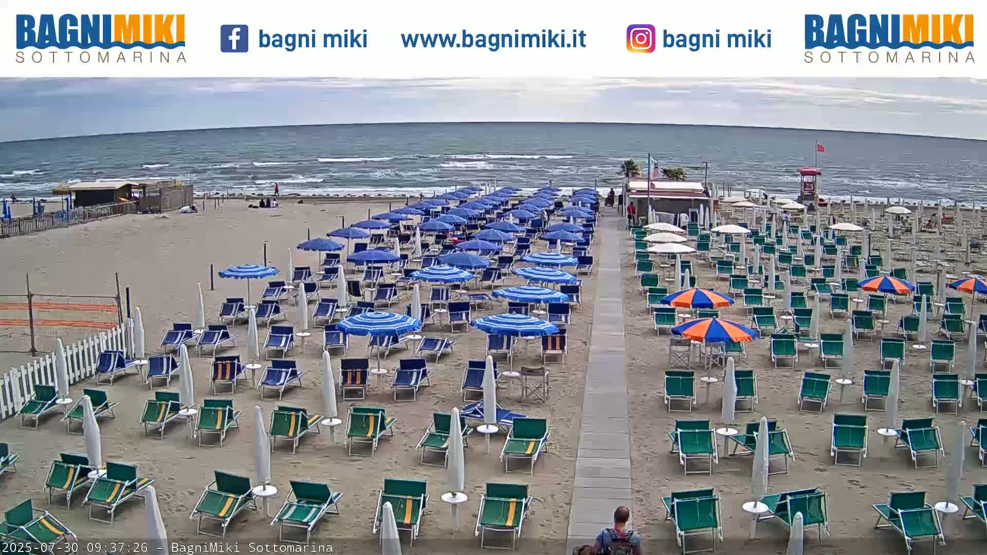 A mostly sunny beach with many rows of beach chairs and umbrellas under an overcast sky, and a calm sea in the background.