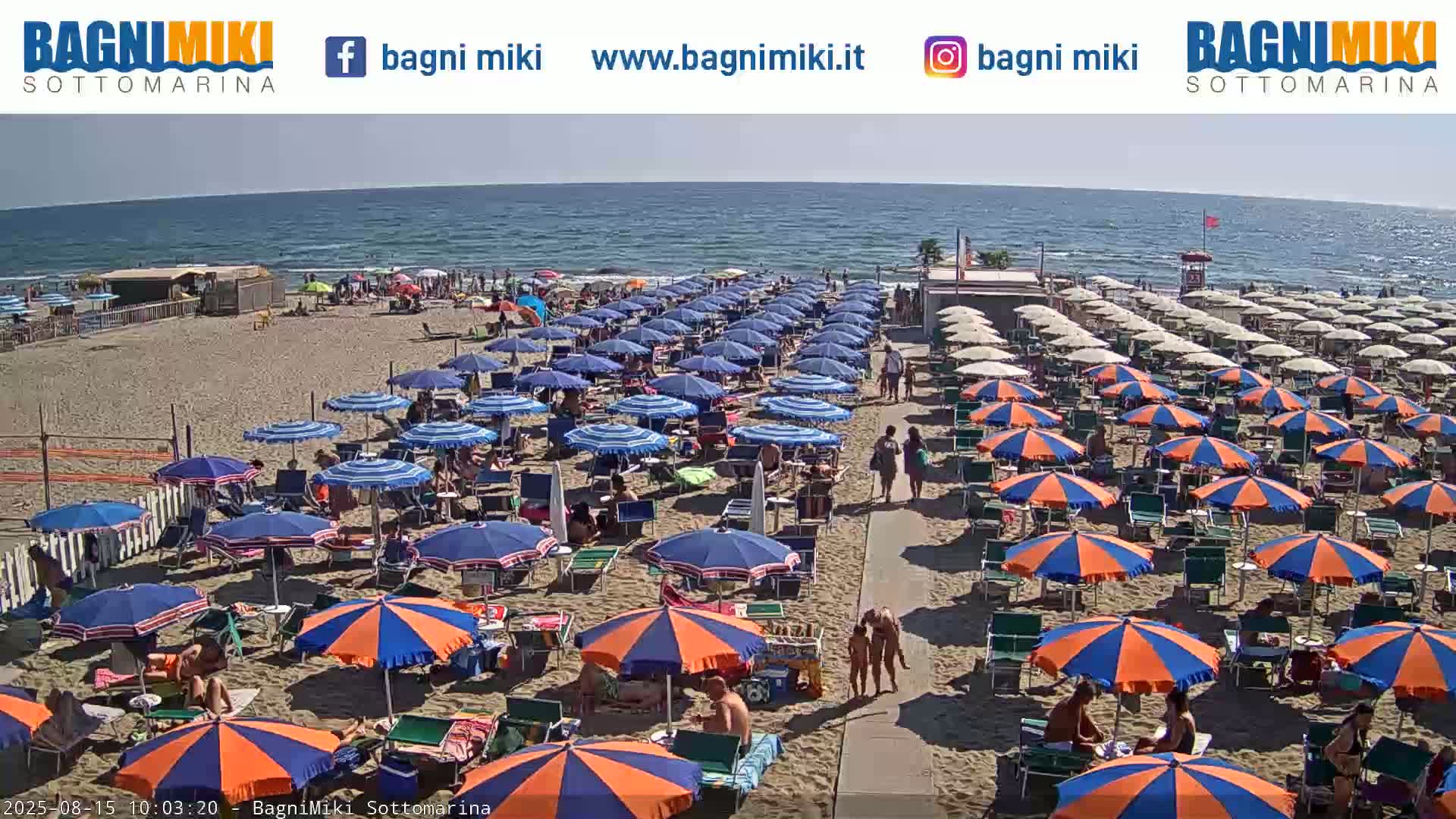 A sunny beach scene shows many blue and orange umbrellas lining a sandy beach next to a calm ocean.