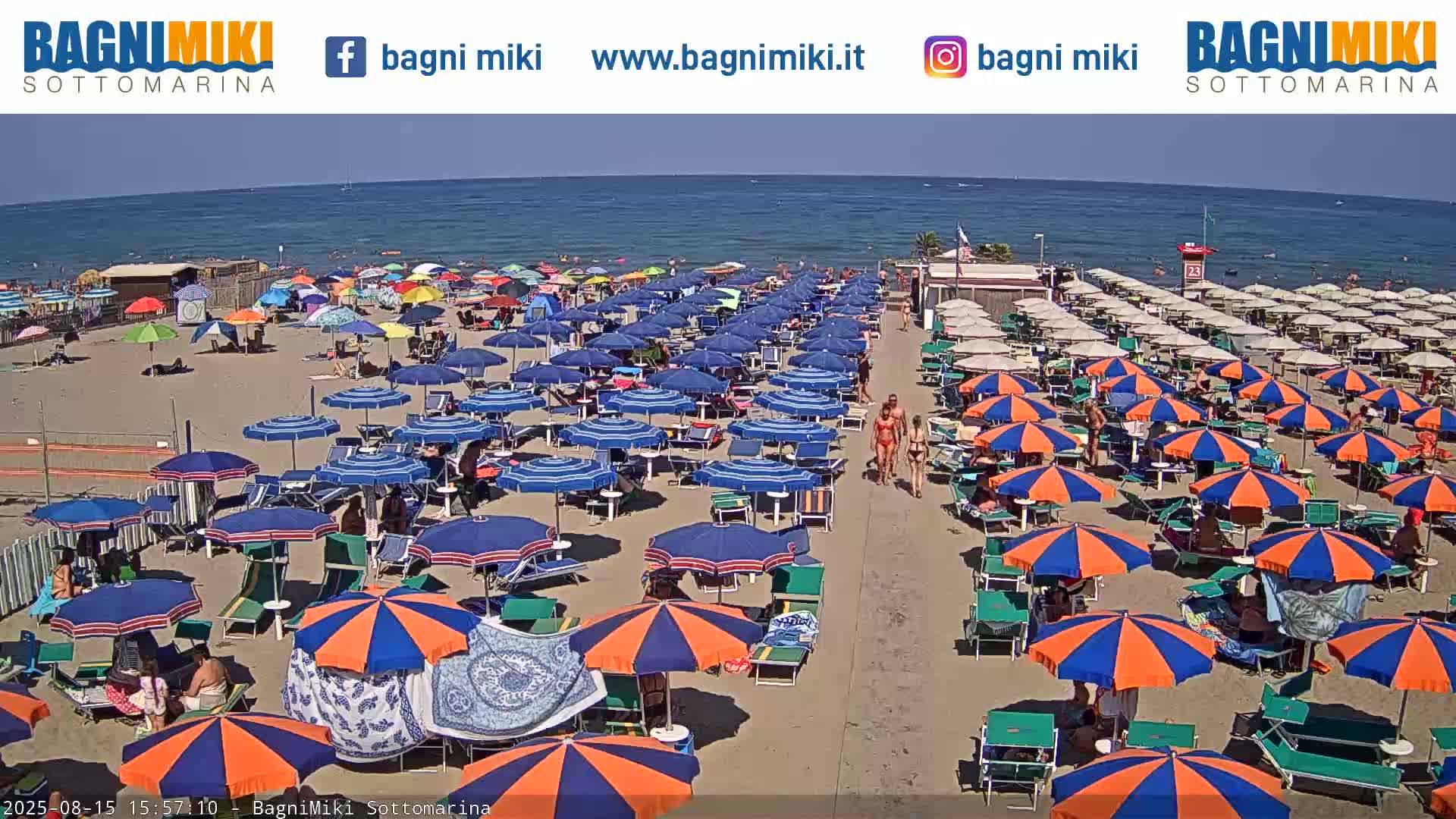 A sunny beach scene shows many blue and orange umbrellas lining a sandy beach next to a calm ocean.