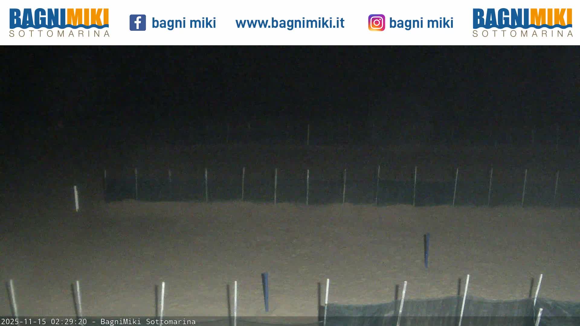 A dimly lit outdoor scene at night displays a sandy ground featuring many thin, mostly light-colored vertical poles, including one distinctly blue, under dark, featureless skies.