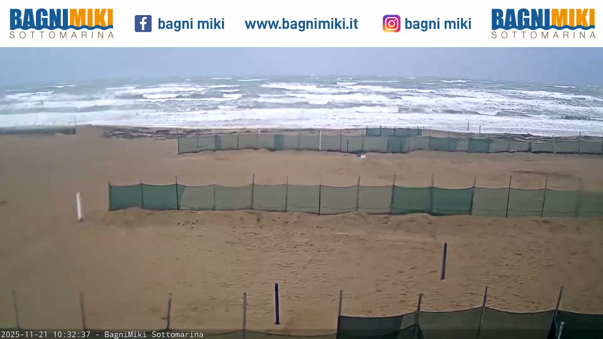 A rough, white-capped sea crashes onto a wide, sandy beach lined with green mesh fencing under an overcast sky.