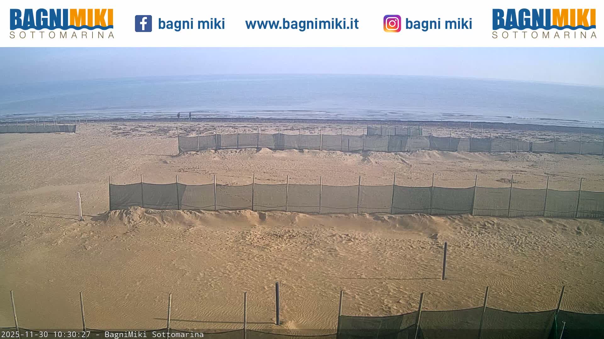 A wide, tan sandy beach with several rows of green sand fences stretches towards a calm, light blue ocean under a hazy, clear sky, with a few distant figures near the water's edge.