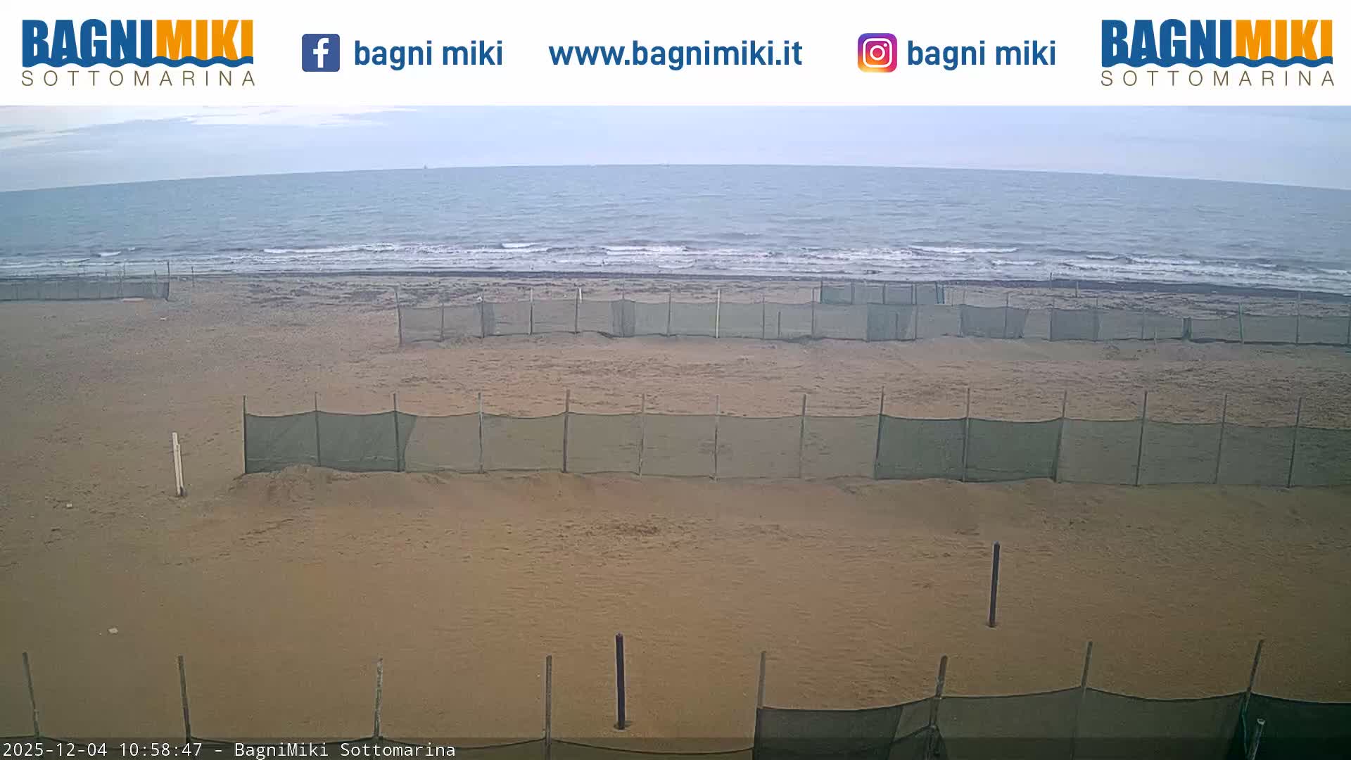 A wide sandy beach is visible with multiple rows of green mesh fencing erected across it, leading to a calm, grey sea under an overcast sky.