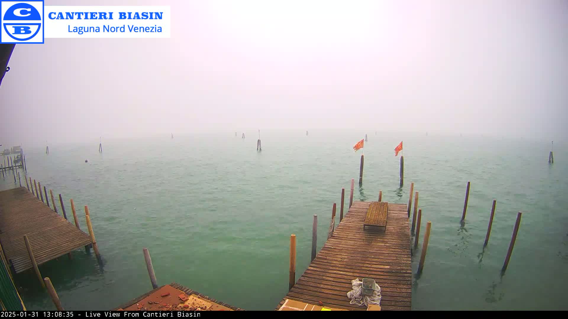 A wooden dock extends over a foggy body of water with several posts and orange flags visible in the distance.