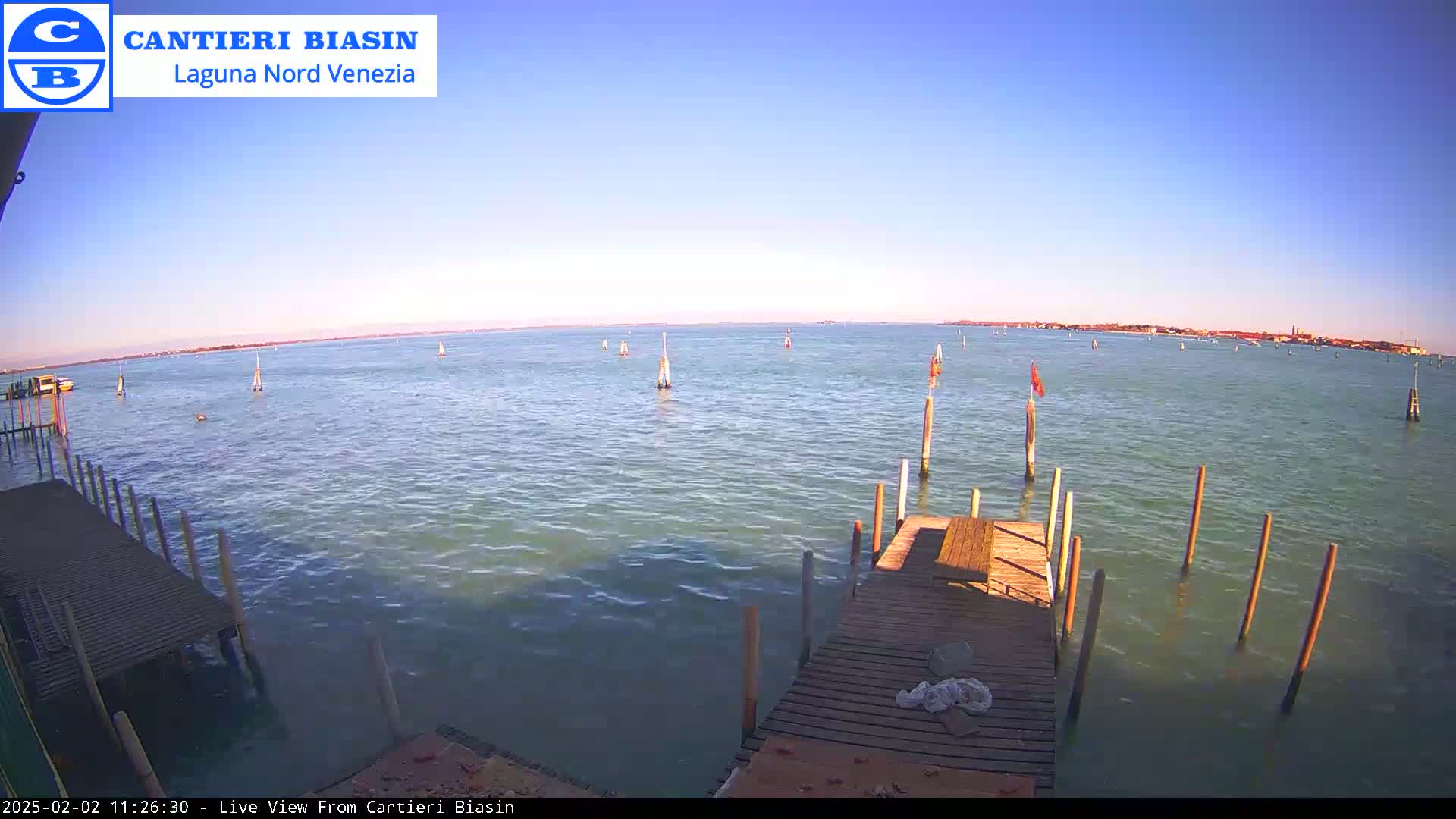 A wooden dock extends over calm, light-blue water on a clear, sunny day, with sailboats and markers visible in the distance.