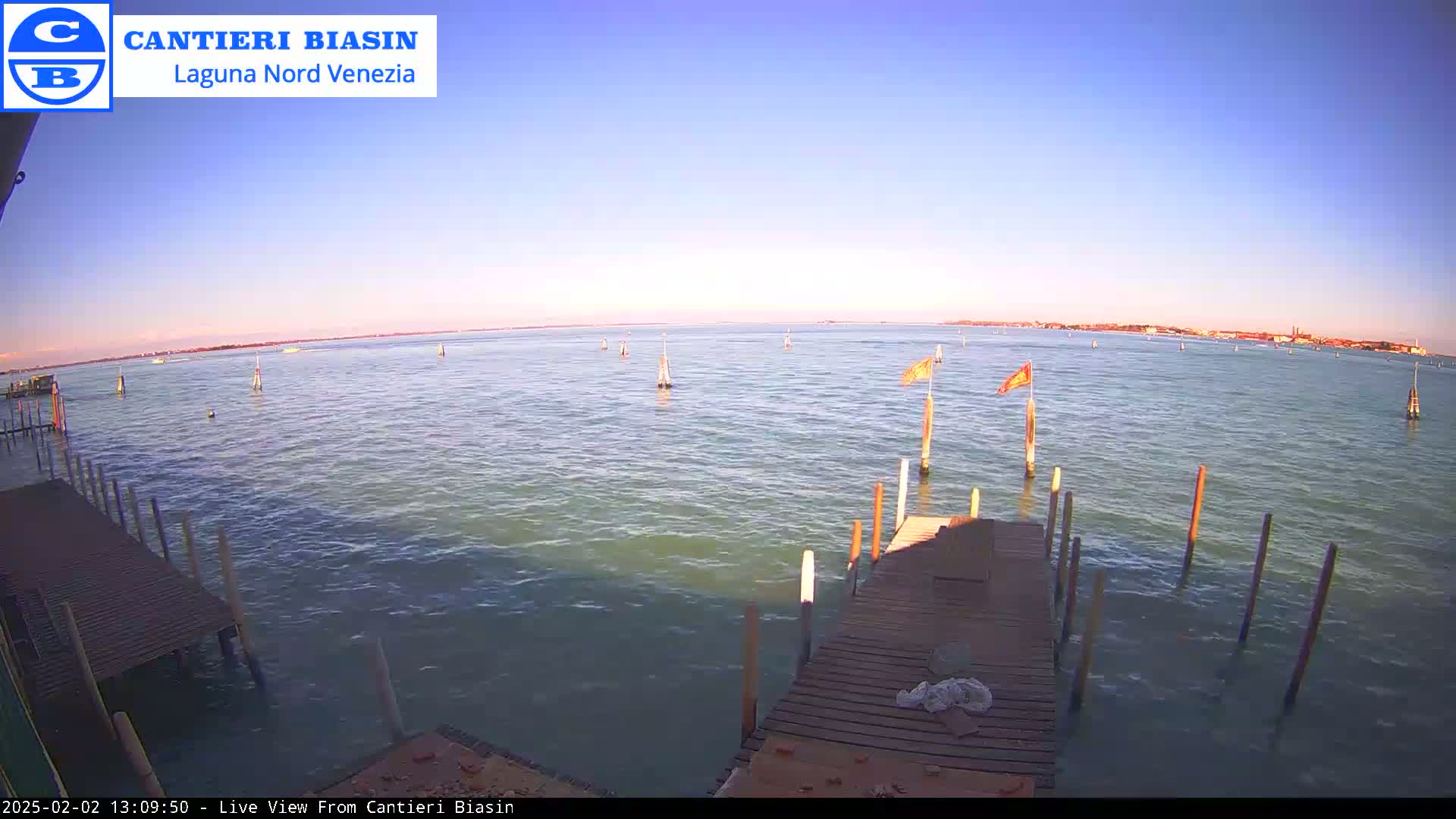 A wooden dock extends over calm, greenish-blue water under a clear, pale-blue sky at sunset, with distant land visible on the horizon.