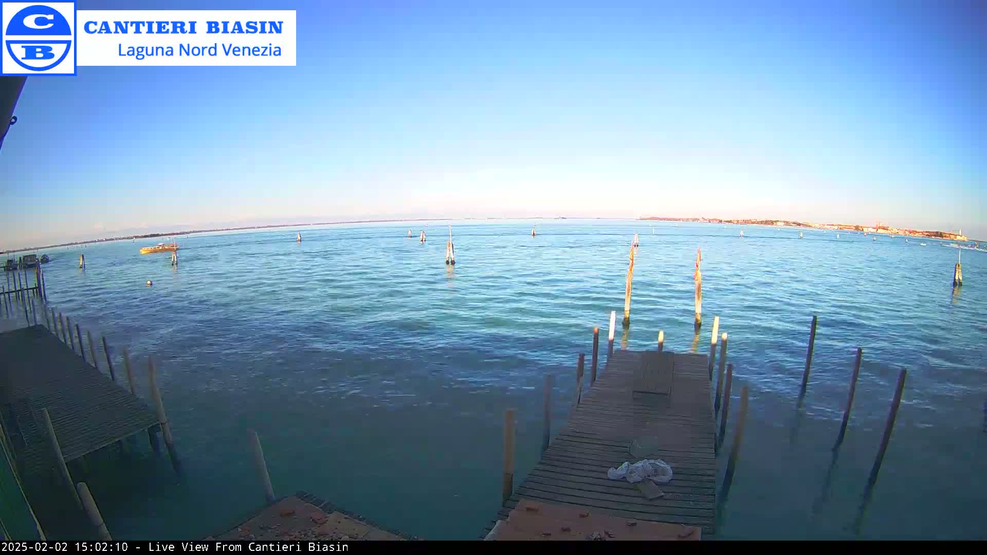 A calm, clear day shows a body of water with several wooden posts extending from a wooden dock, a small boat in the distance, and land visible on the horizon.