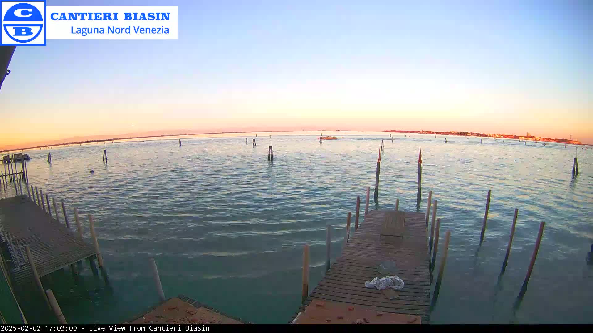 A calm body of water at sunset is viewed from a wooden dock with several posts extending into the water.