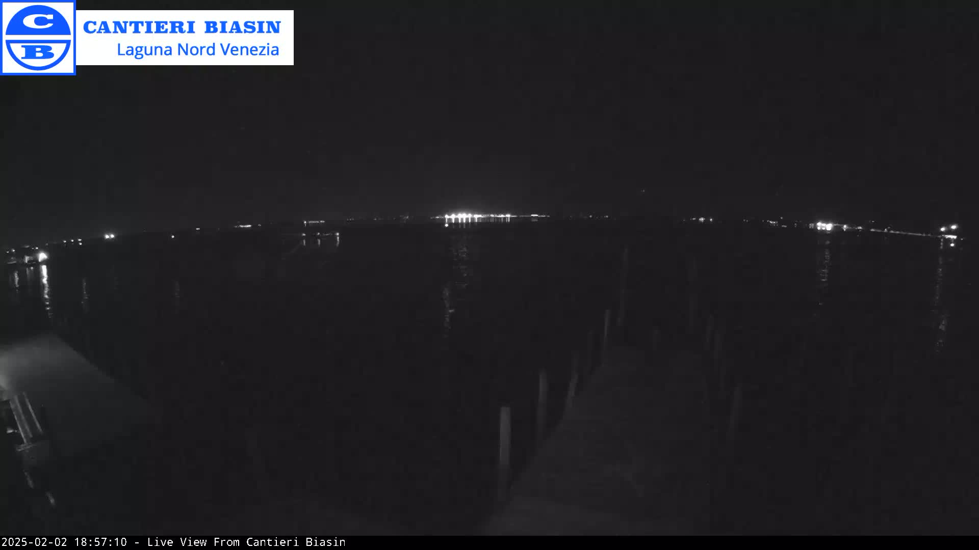 A nighttime view of a dark body of water with distant lights across the horizon and a dark dock in the foreground.
