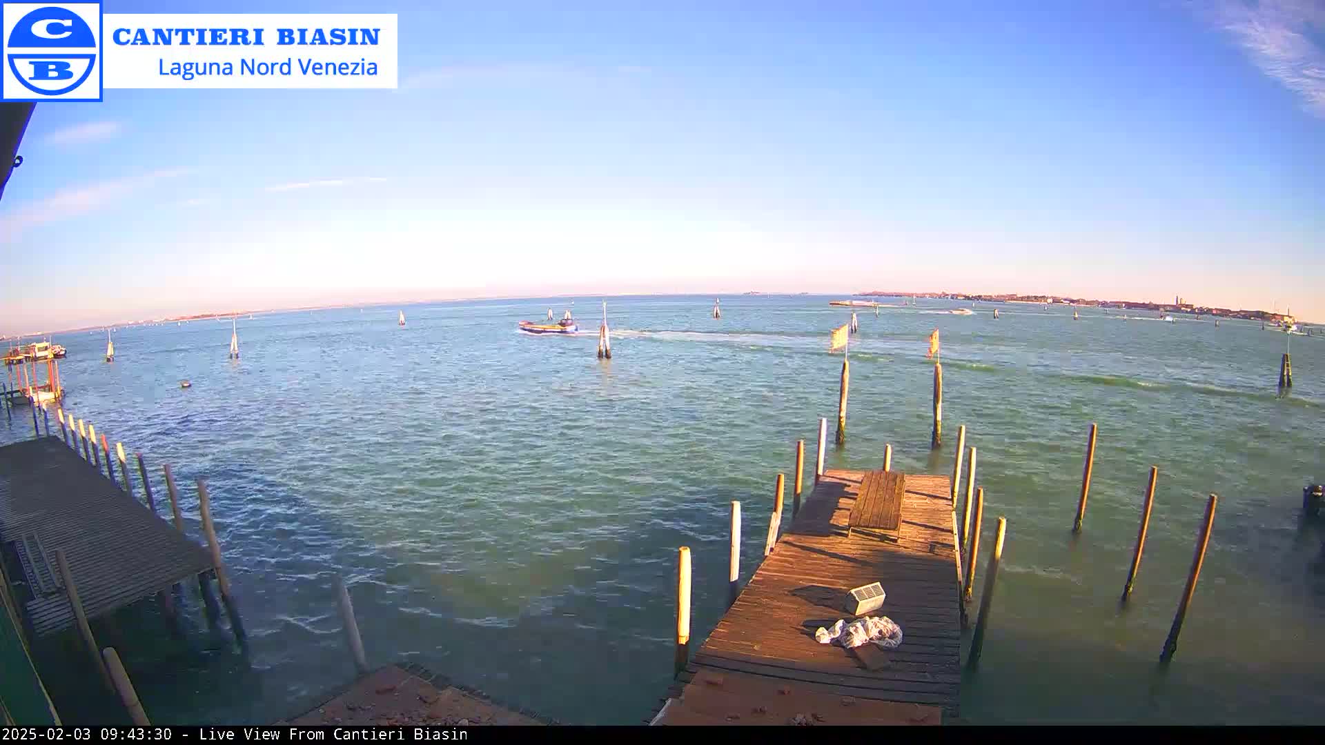 A wooden dock extends over calm, greenish-blue water, with several boats visible in the distance under a clear, bright sky.