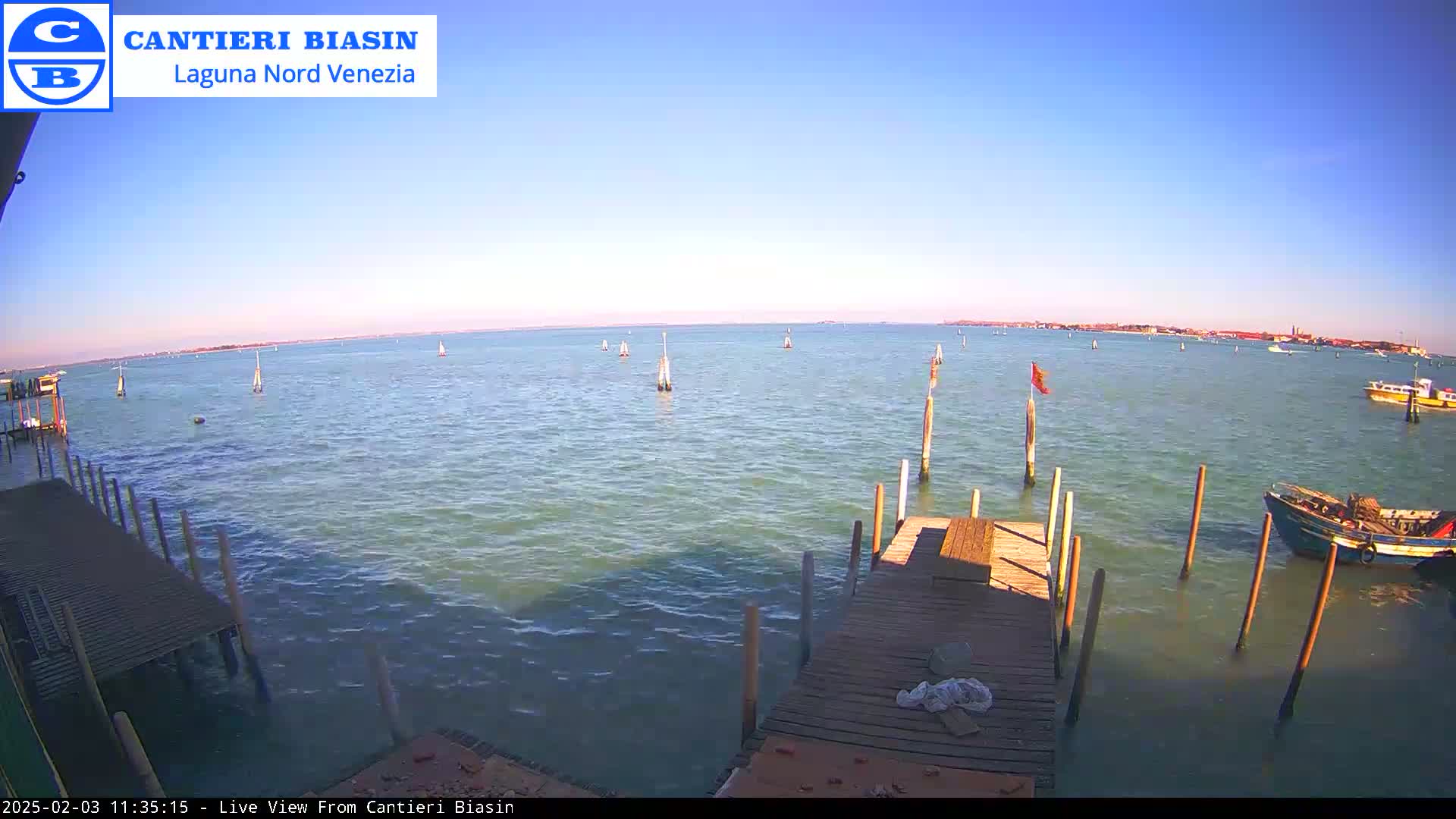 A calm, sunny day shows a wooden dock extending over a body of water with several boats and markers visible in the distance.