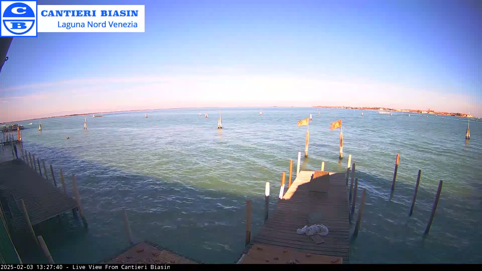 A wooden dock extends over calm, greenish-blue water under a clear, bright blue sky, with a distant shoreline visible.