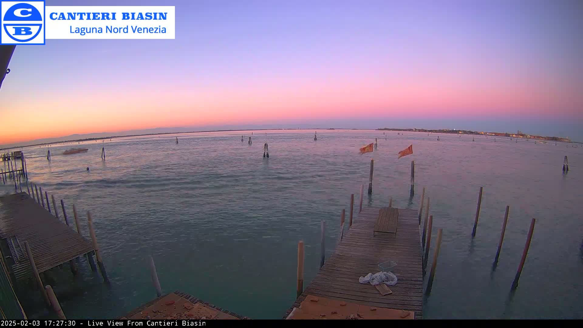 A wooden dock extends over calm water under a pink and purple sunset, with several posts and small flags in the water.