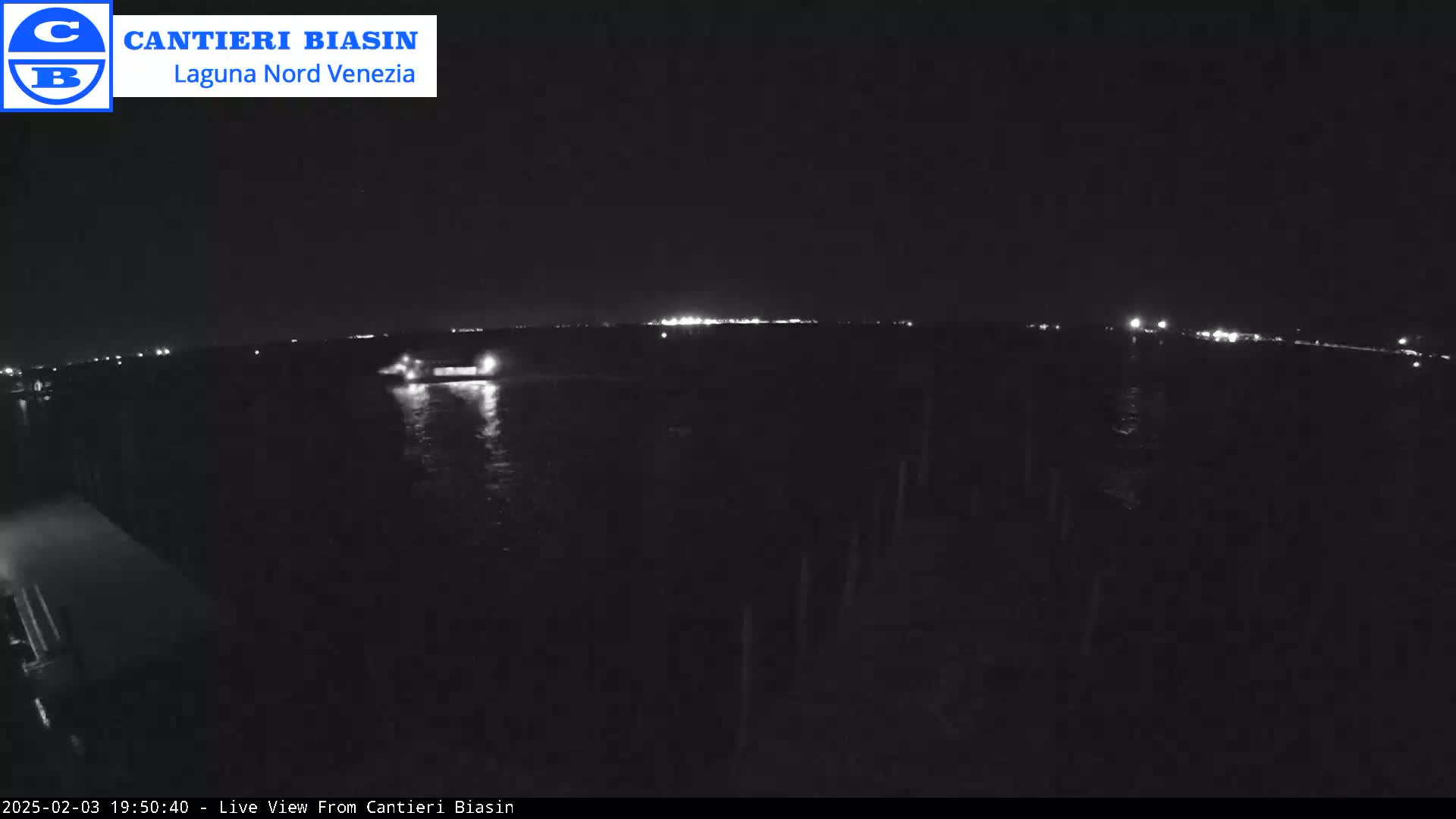 A nighttime, low-light view shows a body of water with a distant shoreline illuminated by lights, and a vessel moving on the water.