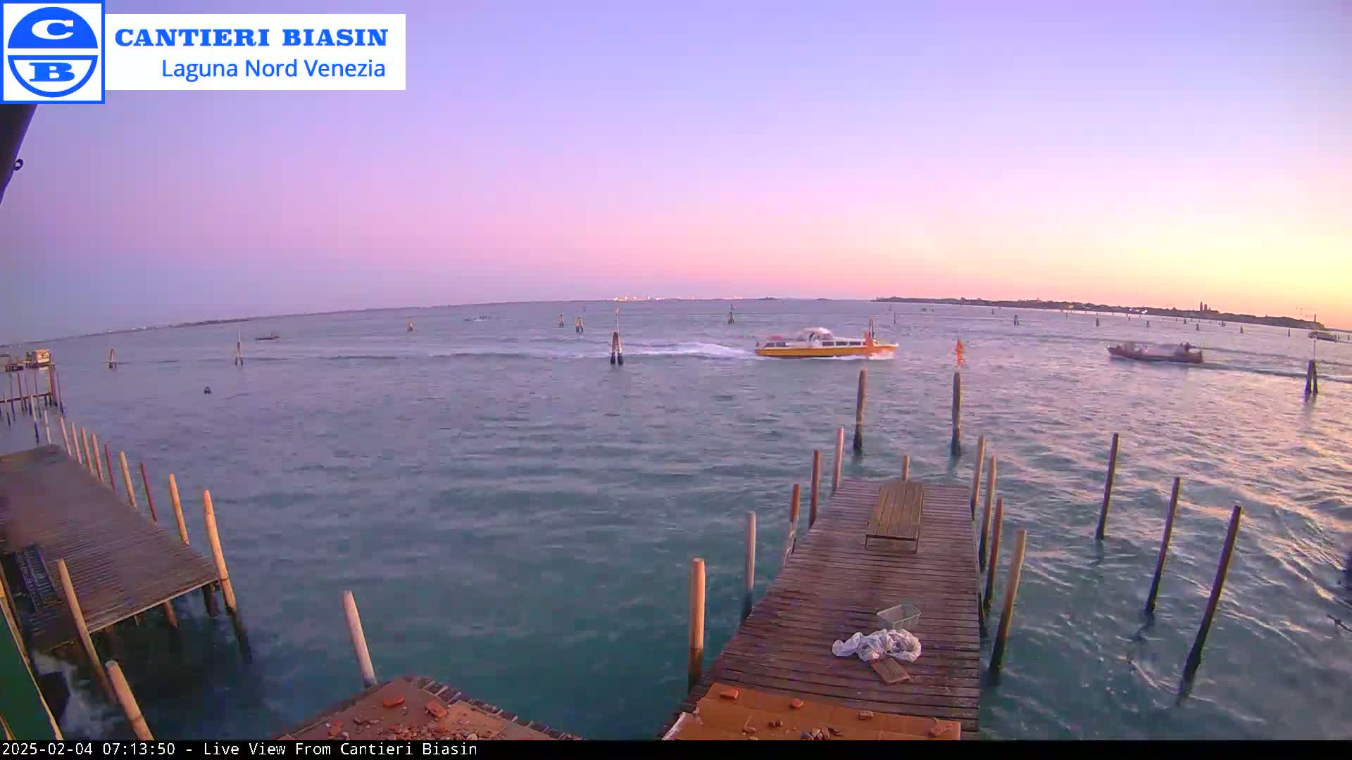 A wooden dock extends over calm water under a pale pink sunset sky, with several boats visible in the distance.