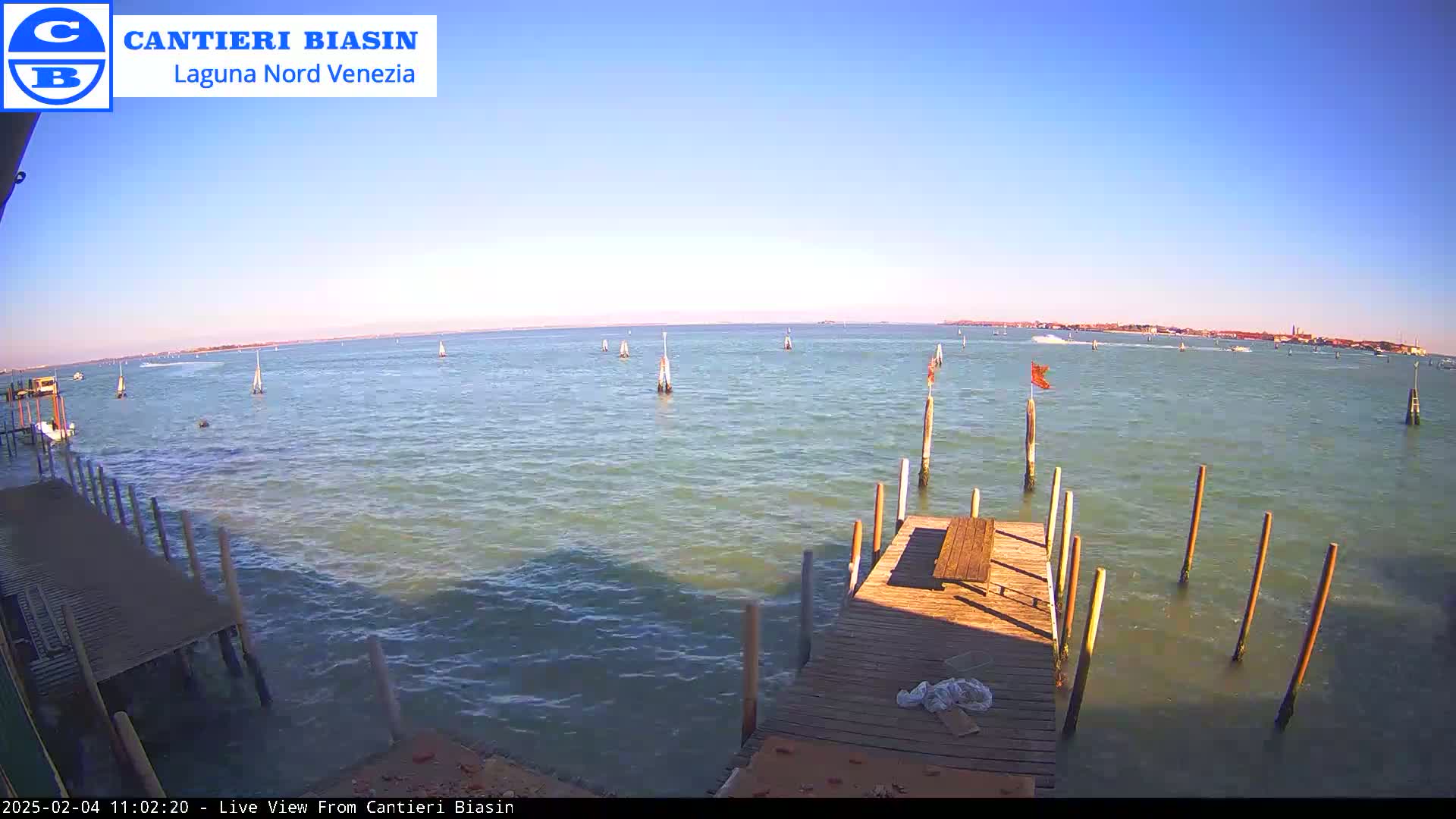 A wooden dock extends over calm, light-green water on a sunny day, with several boats and markers visible in the distance.