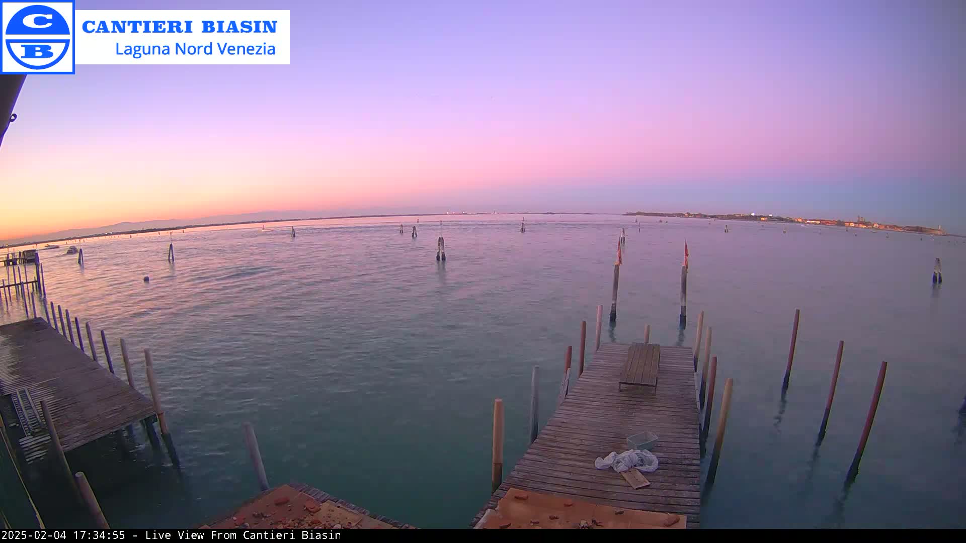 A wooden dock extends over calm, teal water at sunset under a pale pink and purple sky.