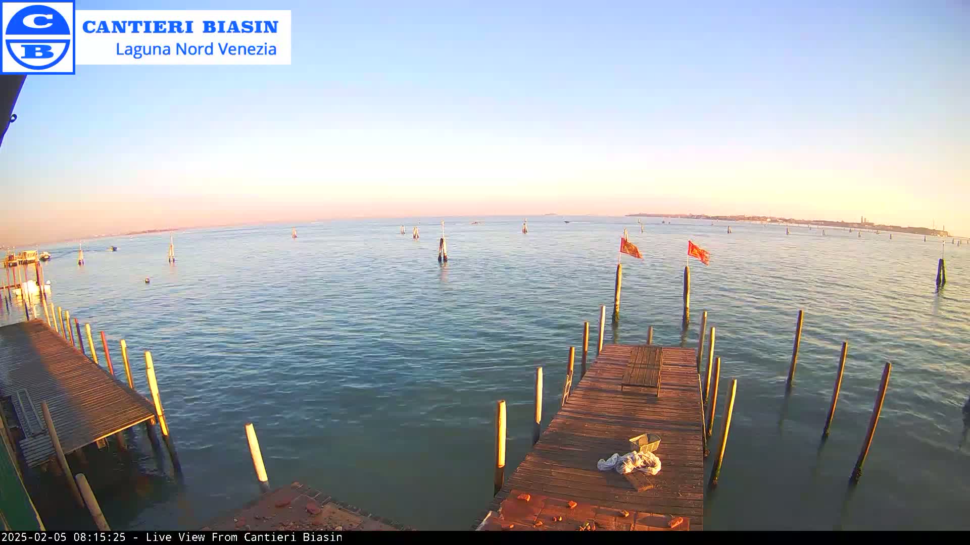 A calm body of water under a clear, light-pink sunset sky is viewed from a wooden dock with several posts extending into the water, some with small flags.