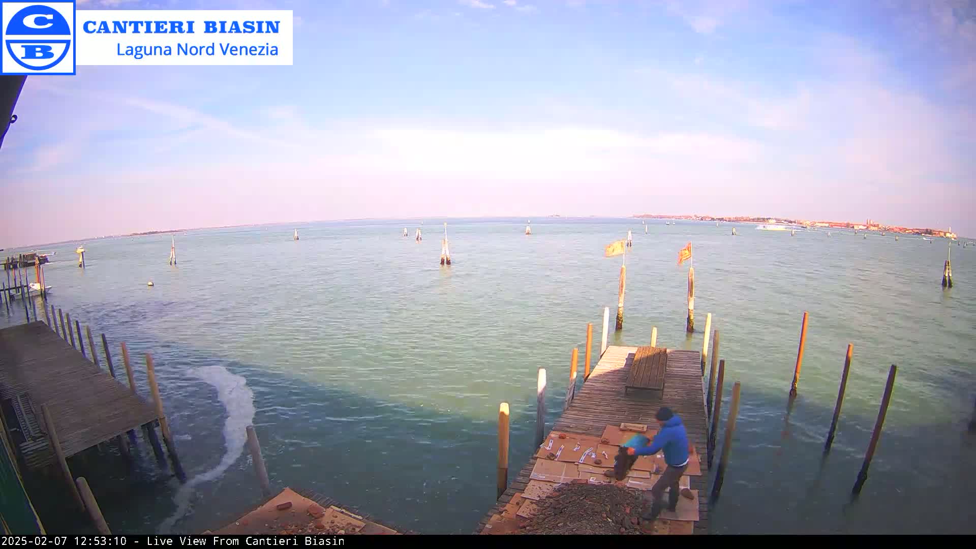 A person works on a wooden dock next to calm, greenish-blue water under a mostly clear sky, with a distant cityscape visible on the horizon.