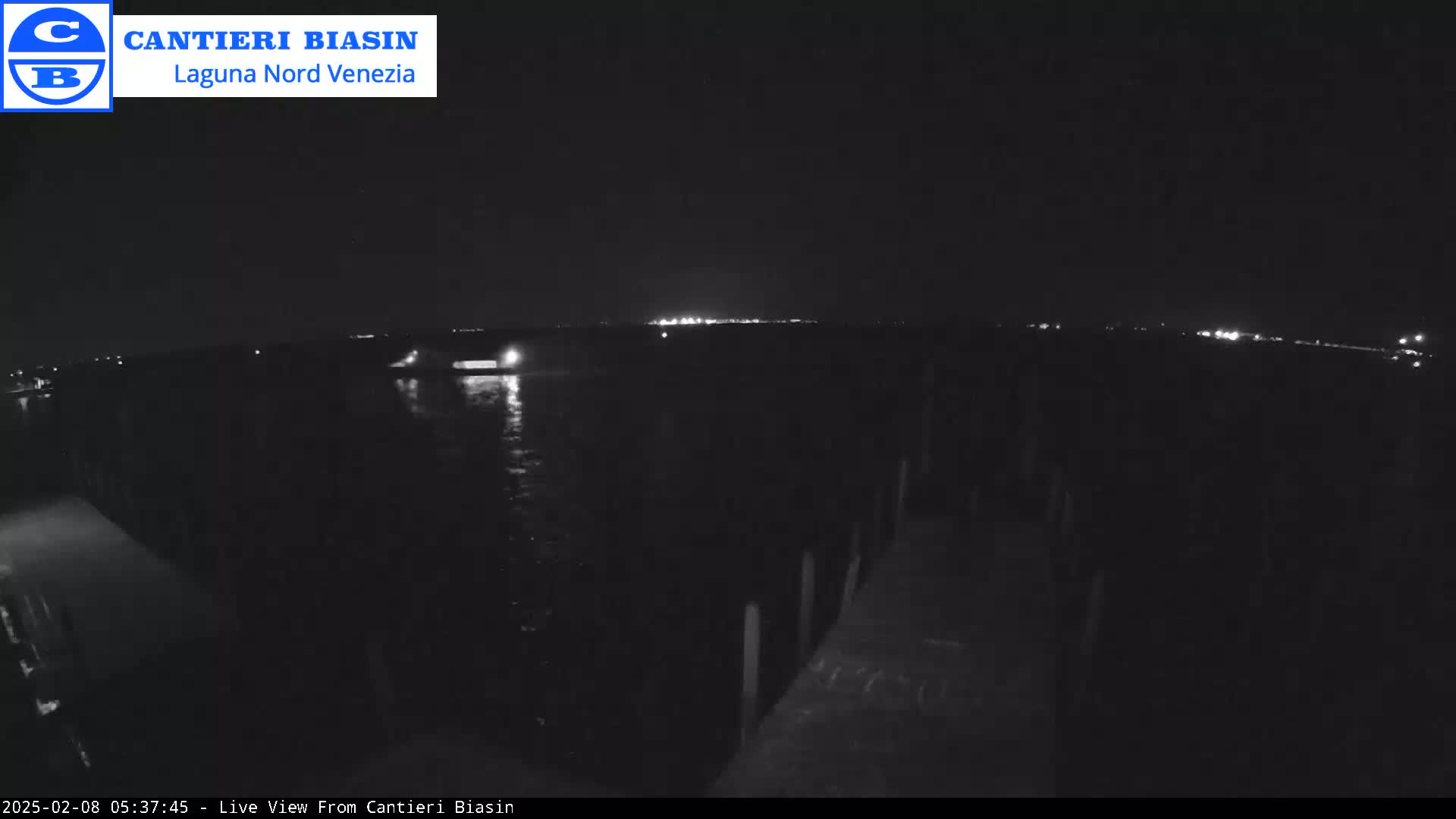A nighttime view of dark water with a distant shoreline showing faint lights, seen from a dark pier or dock.