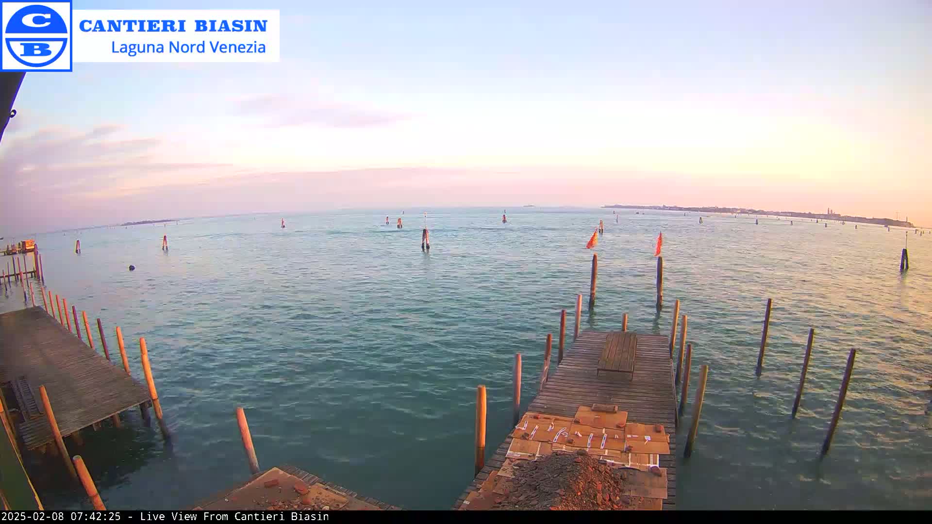 A calm body of water with numerous posts and a wooden dock in the foreground, under a pale pink and orange sunrise sky.