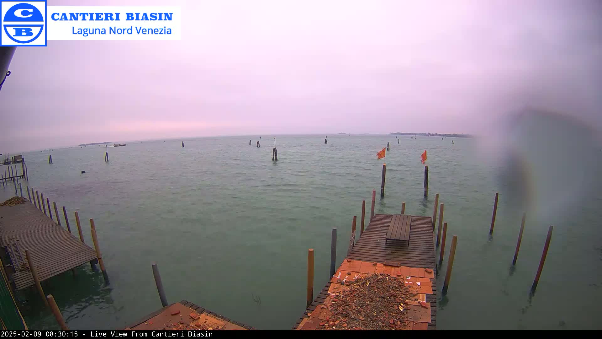 A calm body of water with several wooden pilings and a small dock is visible under an overcast sky.