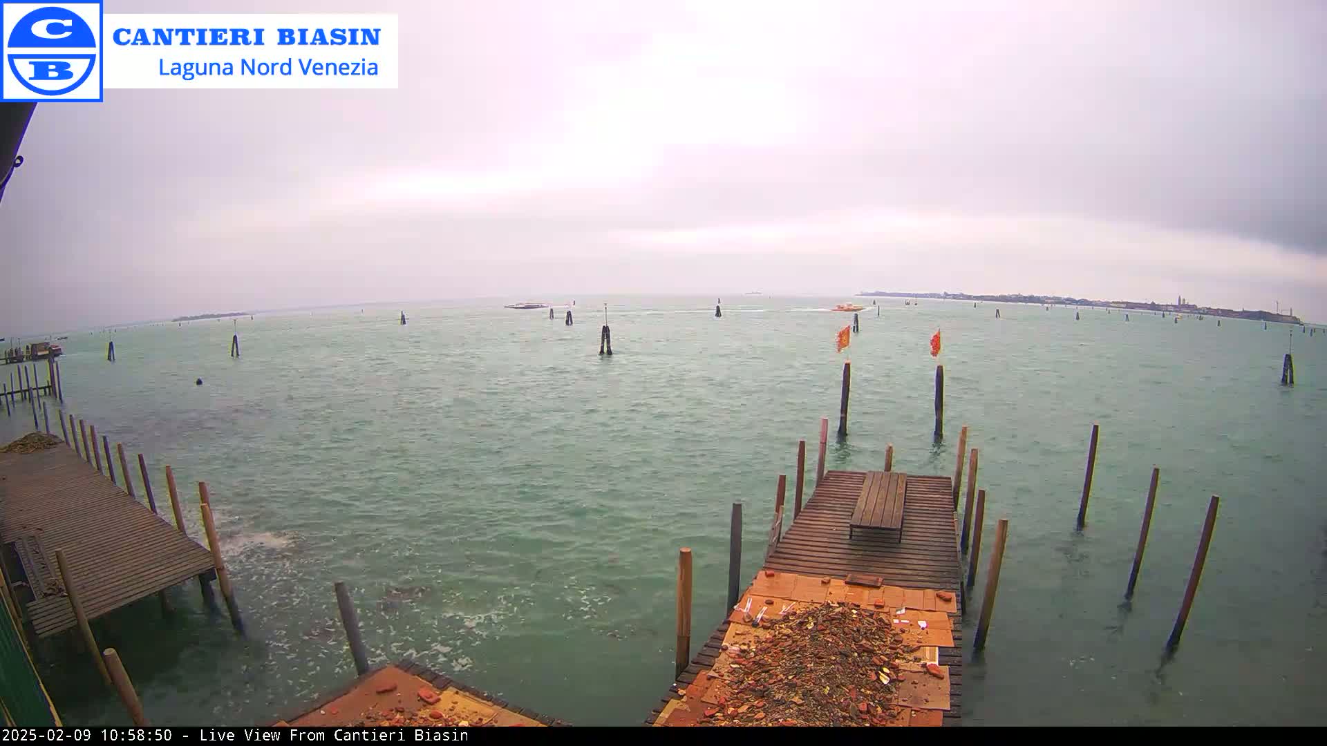 A view of a calm, overcast body of water with several wooden pilings and a small dock in the foreground, a few boats in the distance, and land visible on the horizon.
