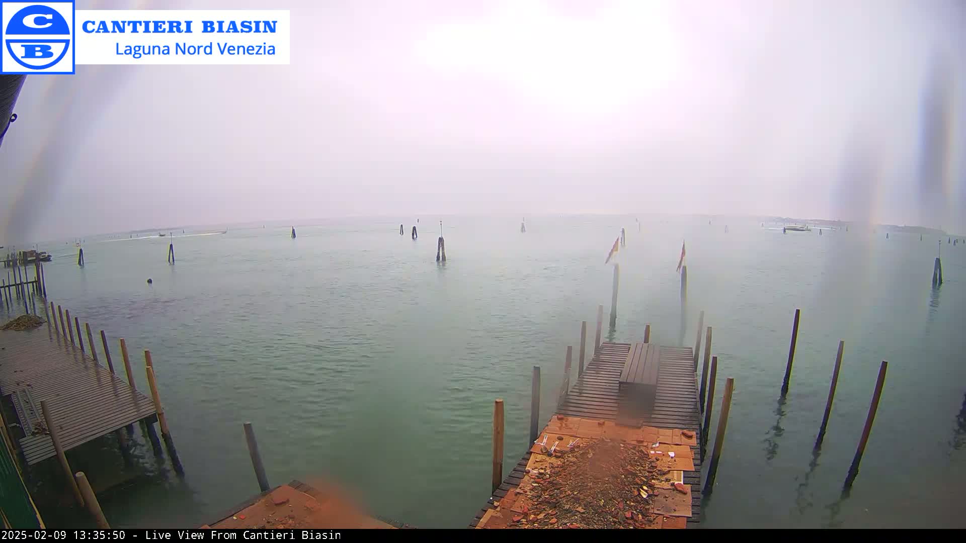 A hazy, overcast day reveals a calm body of water with wooden pilings and a small dock, showing a few boats in the distance.