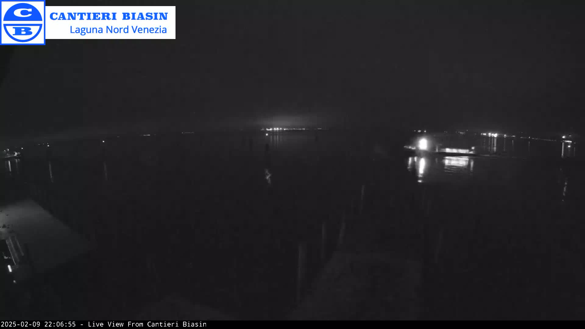 A nighttime, low-light view of a dark body of water with faint lights visible on the far shore and a few lights closer to the camera.