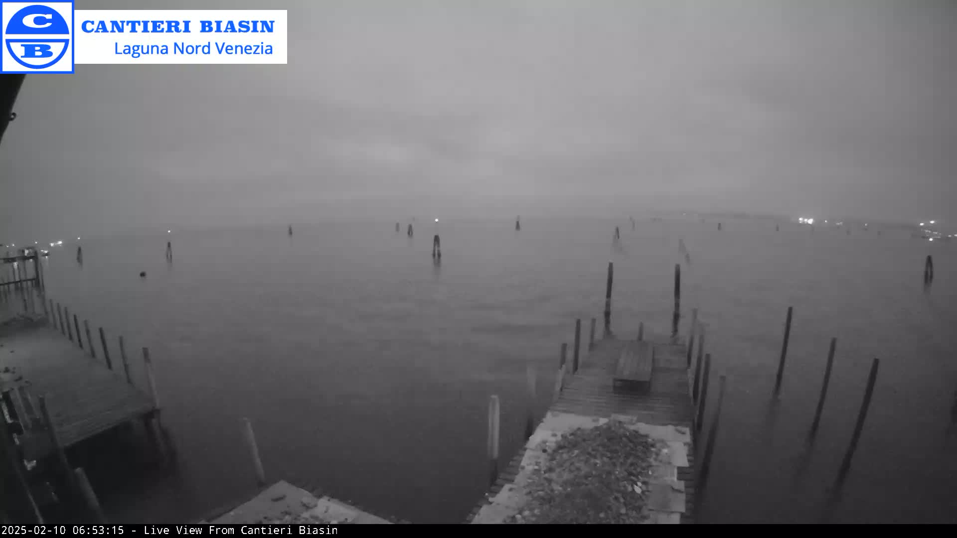 A foggy, gray morning shows a calm body of water with several wooden posts and a dock in the foreground.