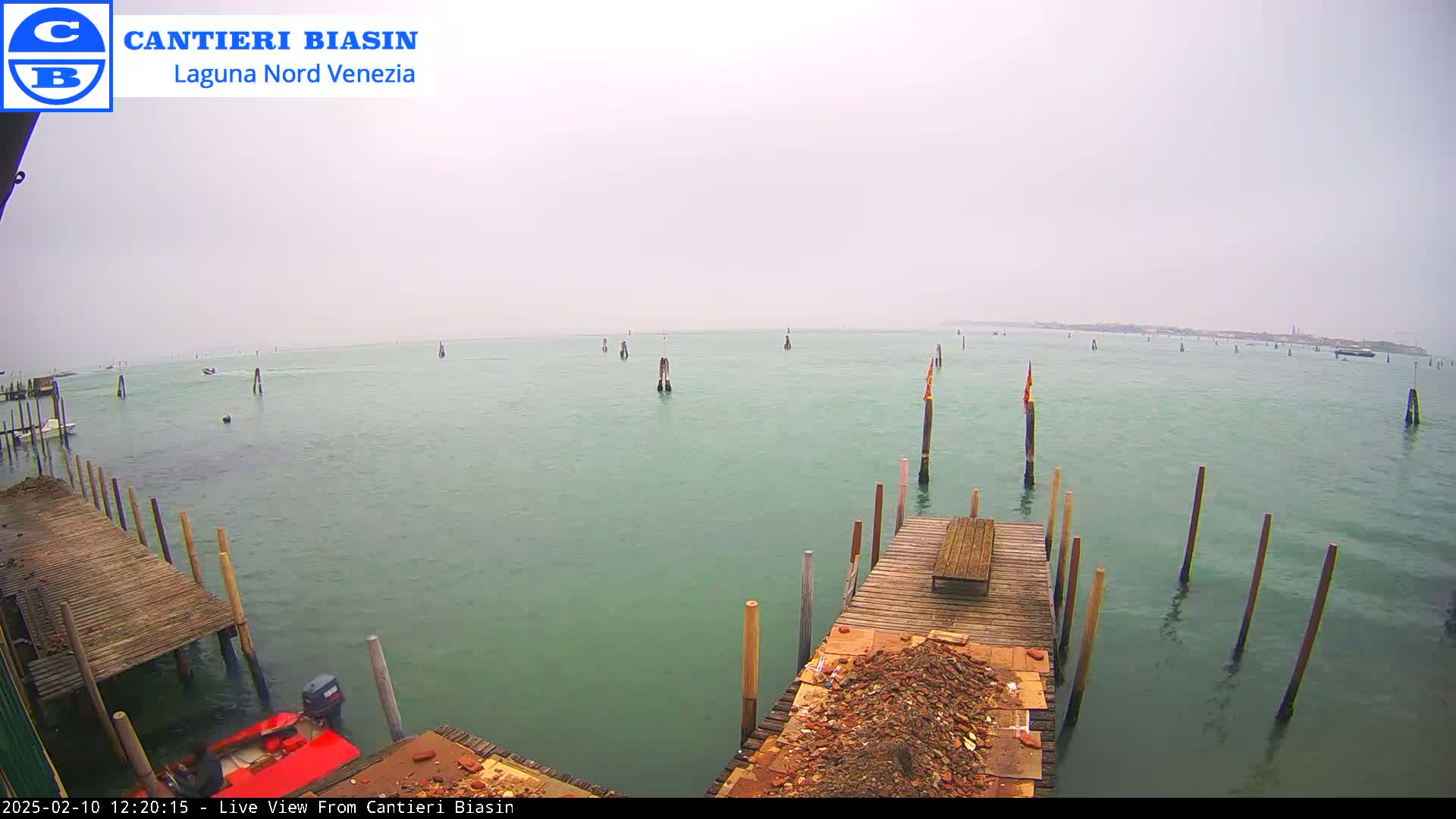A hazy, overcast day shows a body of water with numerous wooden posts and a small red boat near a wooden dock piled with debris.