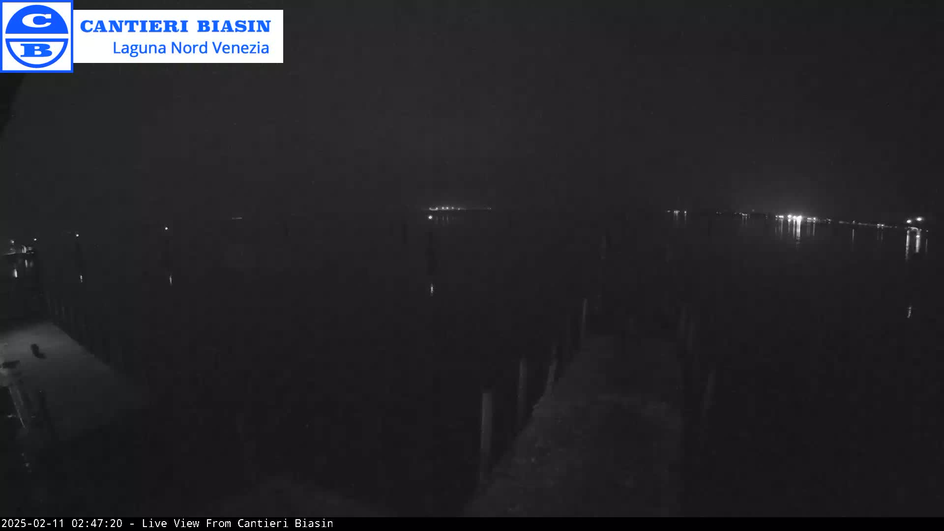 A nighttime view of a dark body of water with faintly visible lights across it and a dock in the foreground.