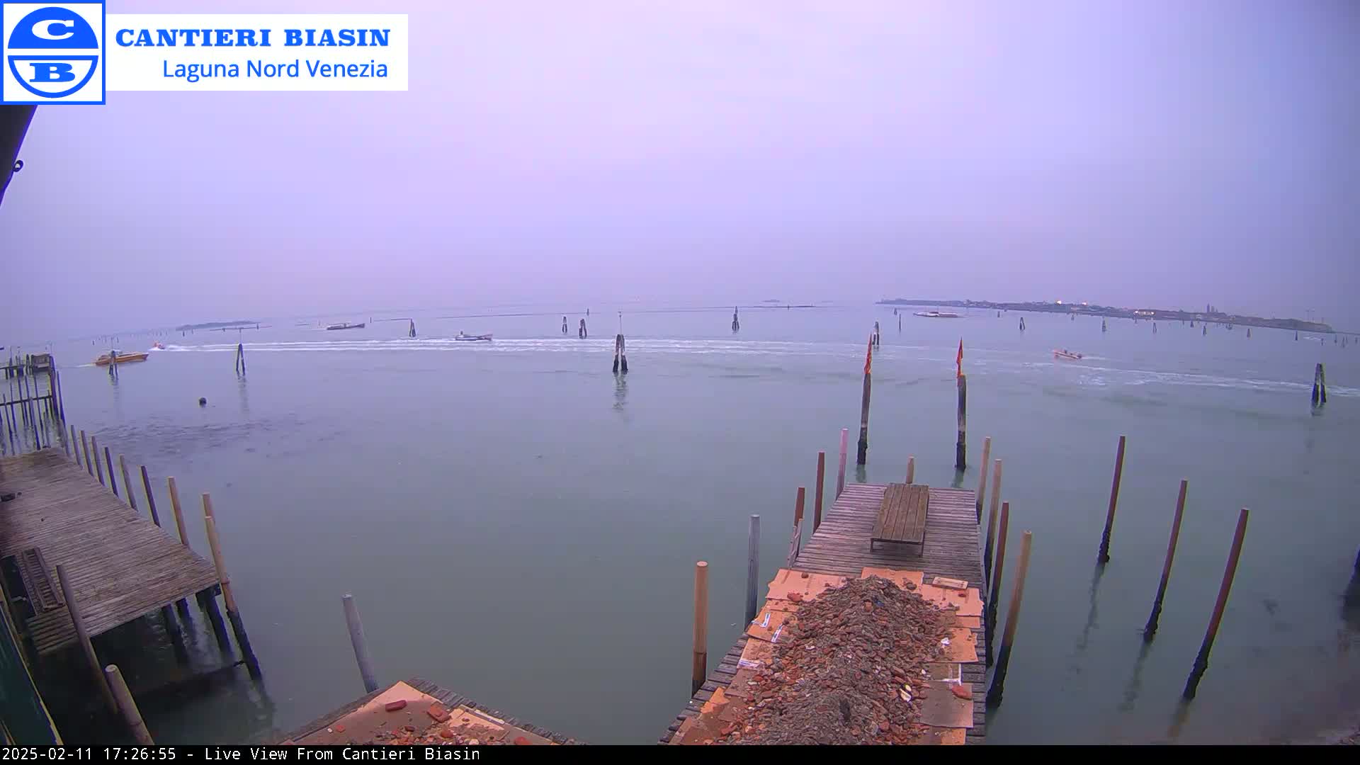 A calm, overcast day reveals a body of water with several boats and wooden pilings, alongside a weathered wooden dock with construction debris.